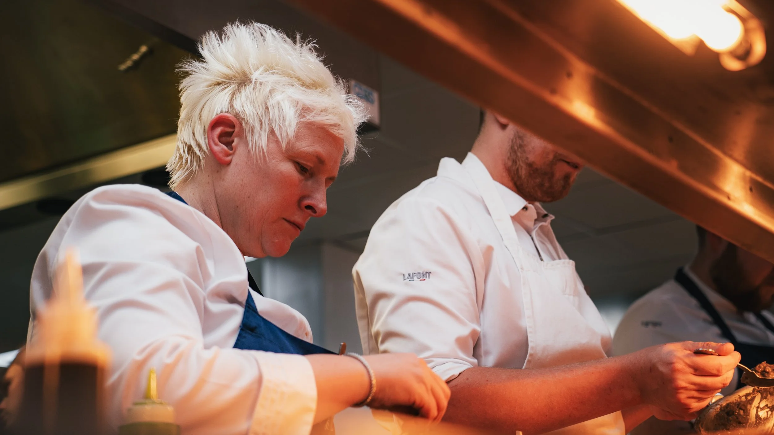 Two chefs wearing white uniforms and aprons preparing food in a professional kitchen, focusing on their work.