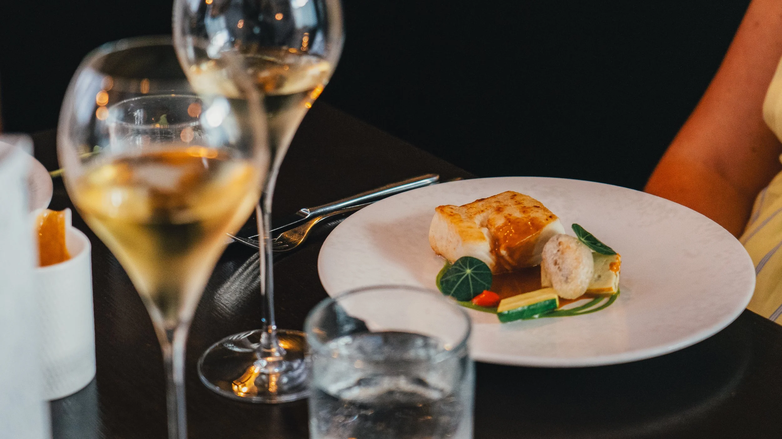 A close-up of a dinner table with a glass of white wine, a plate with a piece of cheesecake garnished with green leaves, and a fork, with other drink glasses and a candle in the background.