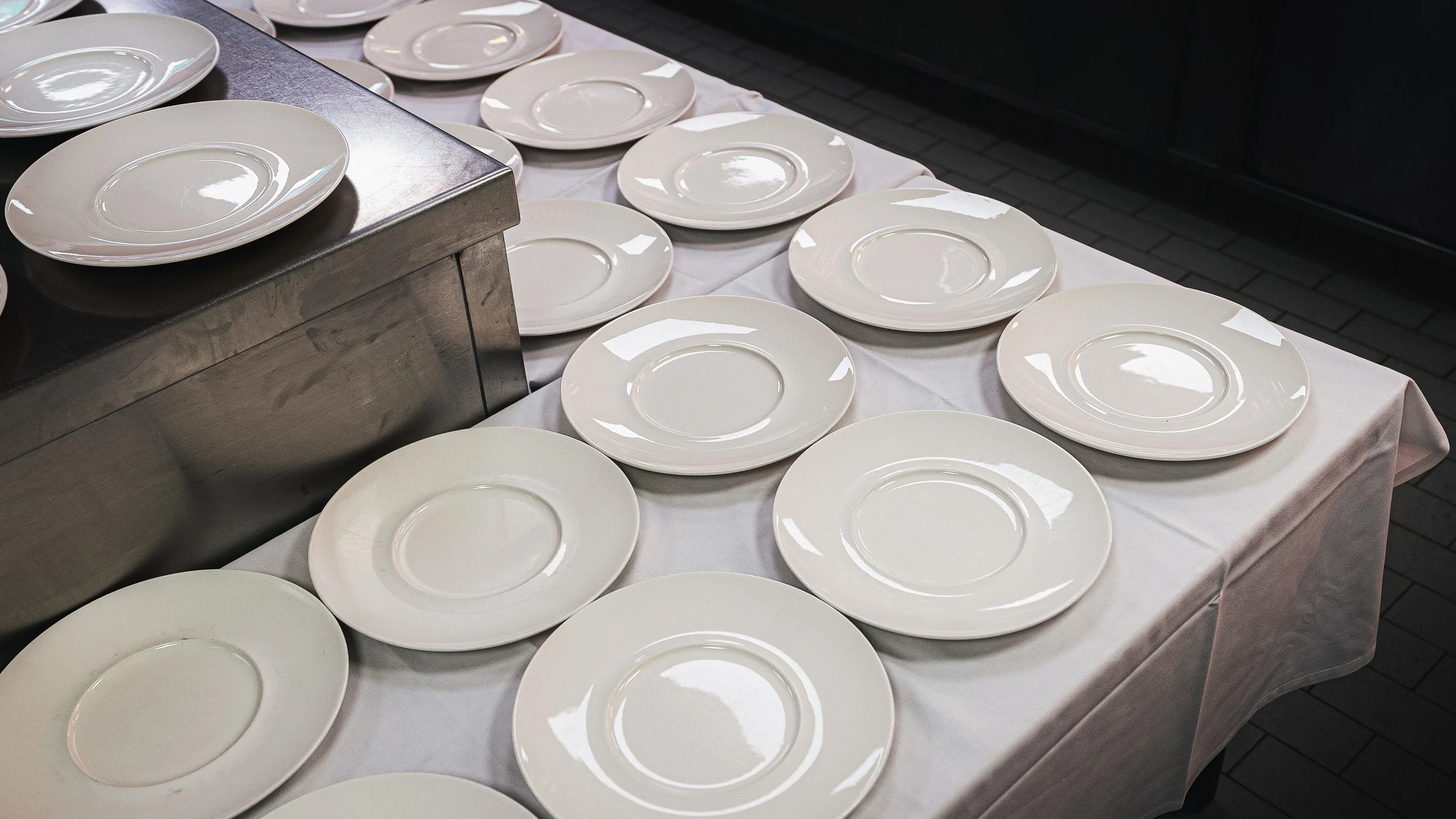 Multiple white plates arranged on a table covered with a white tablecloth. The plates are stacked and spread out, with some on a metal surface. The setting appears to be prepared for serving food.