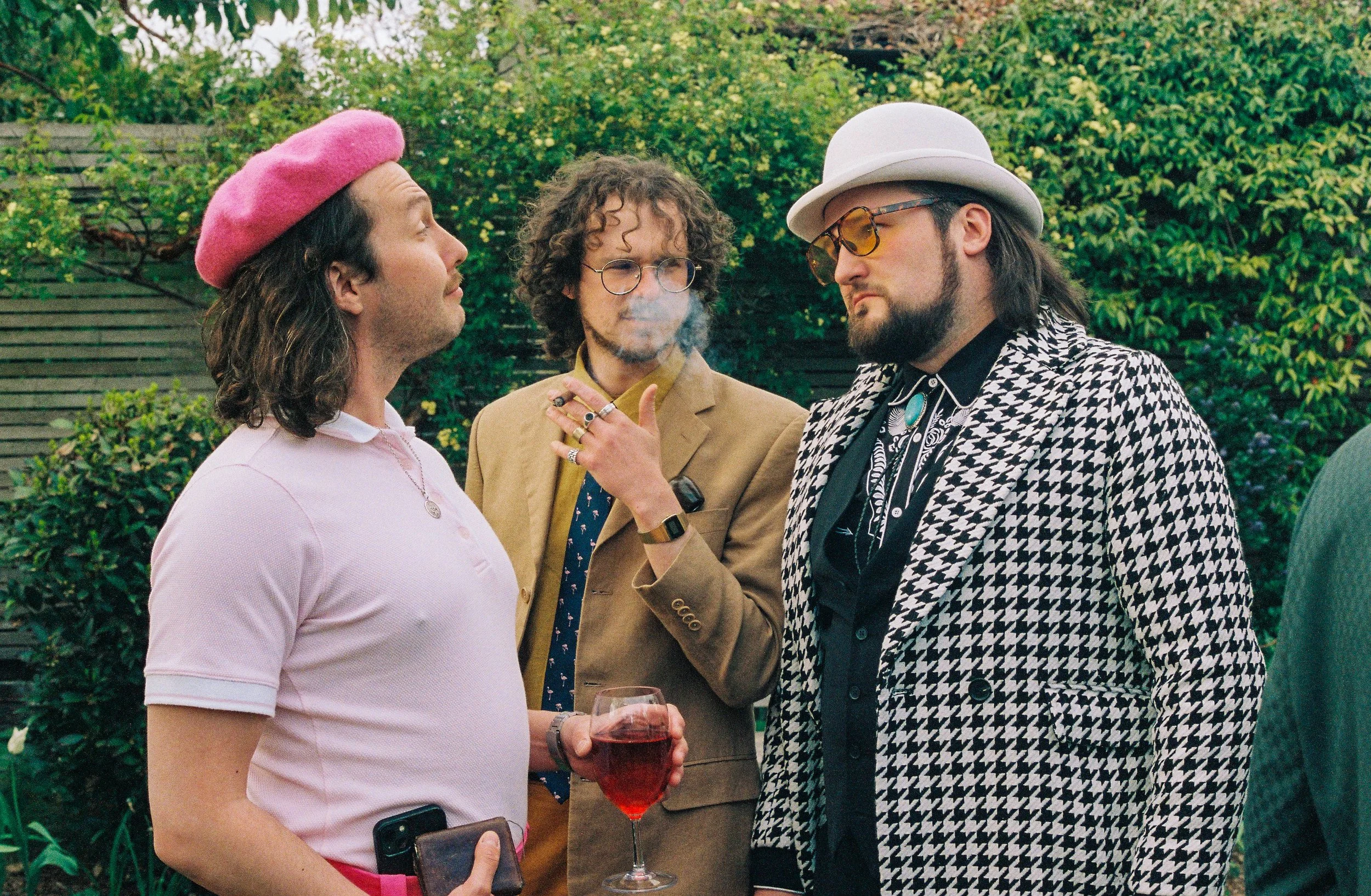 Three men at a social gathering outdoors, one holding a glass of red wine, wearing stylish clothing and accessories, with a background of green foliage.