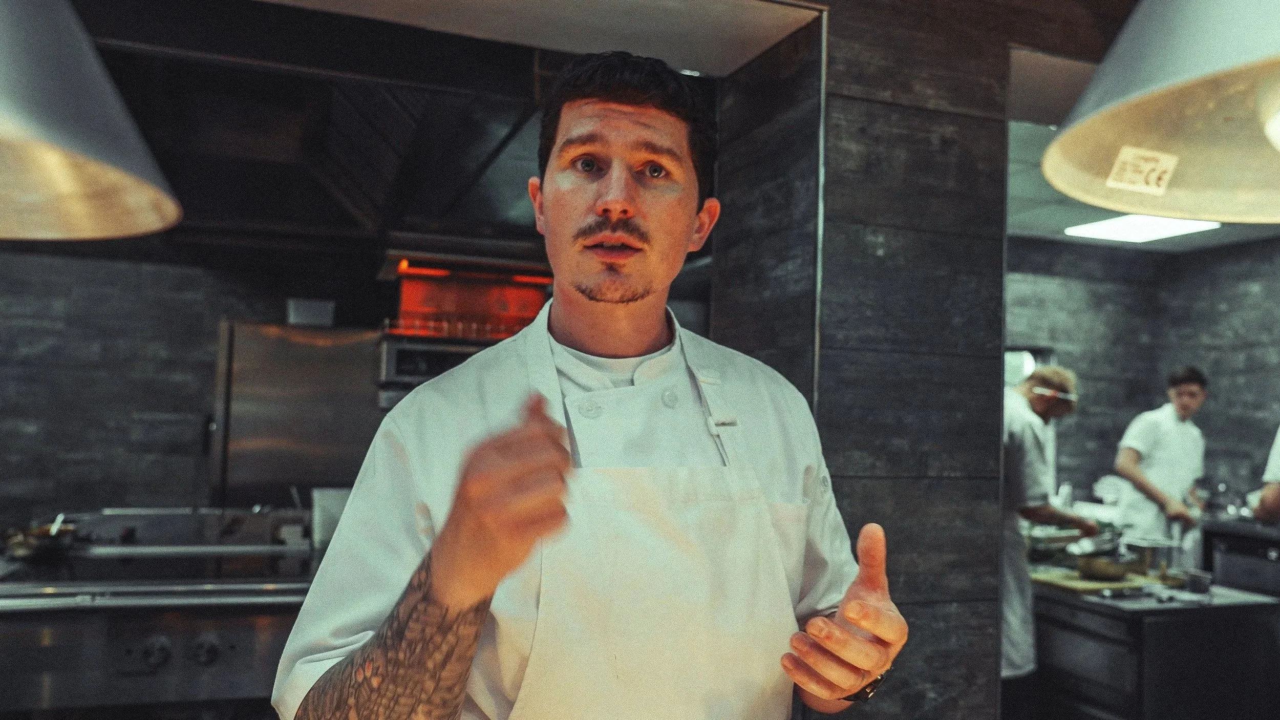 A male chef with dark hair, a mustache, and tattooed arm, wearing a white chef's coat and apron, giving a thumbs-up gesture in a professional kitchen with other staff in the background.
