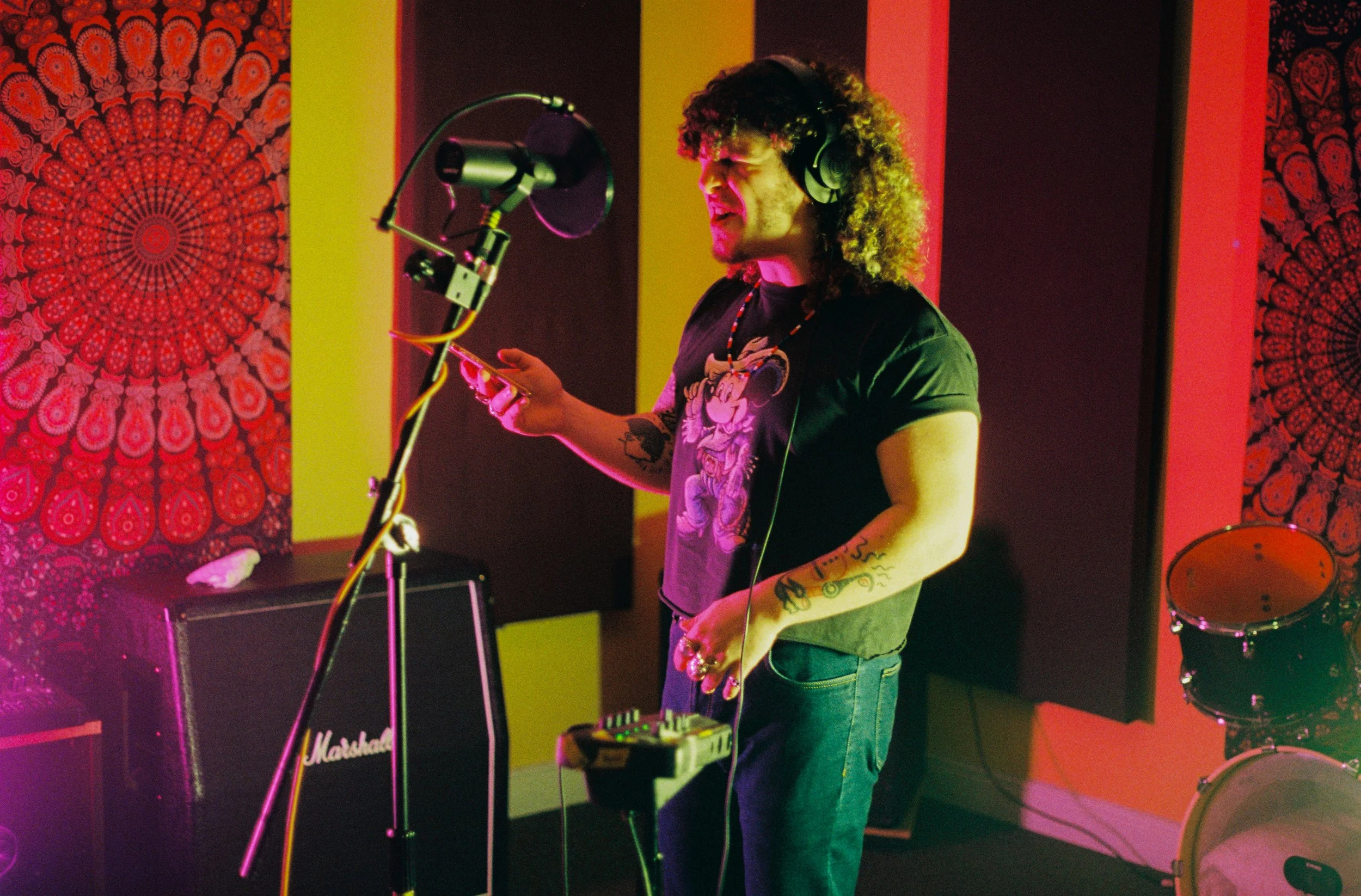 A man with curly hair and tattoos singing into a microphone in a music studio, wearing headphones and playing a keyboard, with colorful psychedelic wall art and musical equipment around.