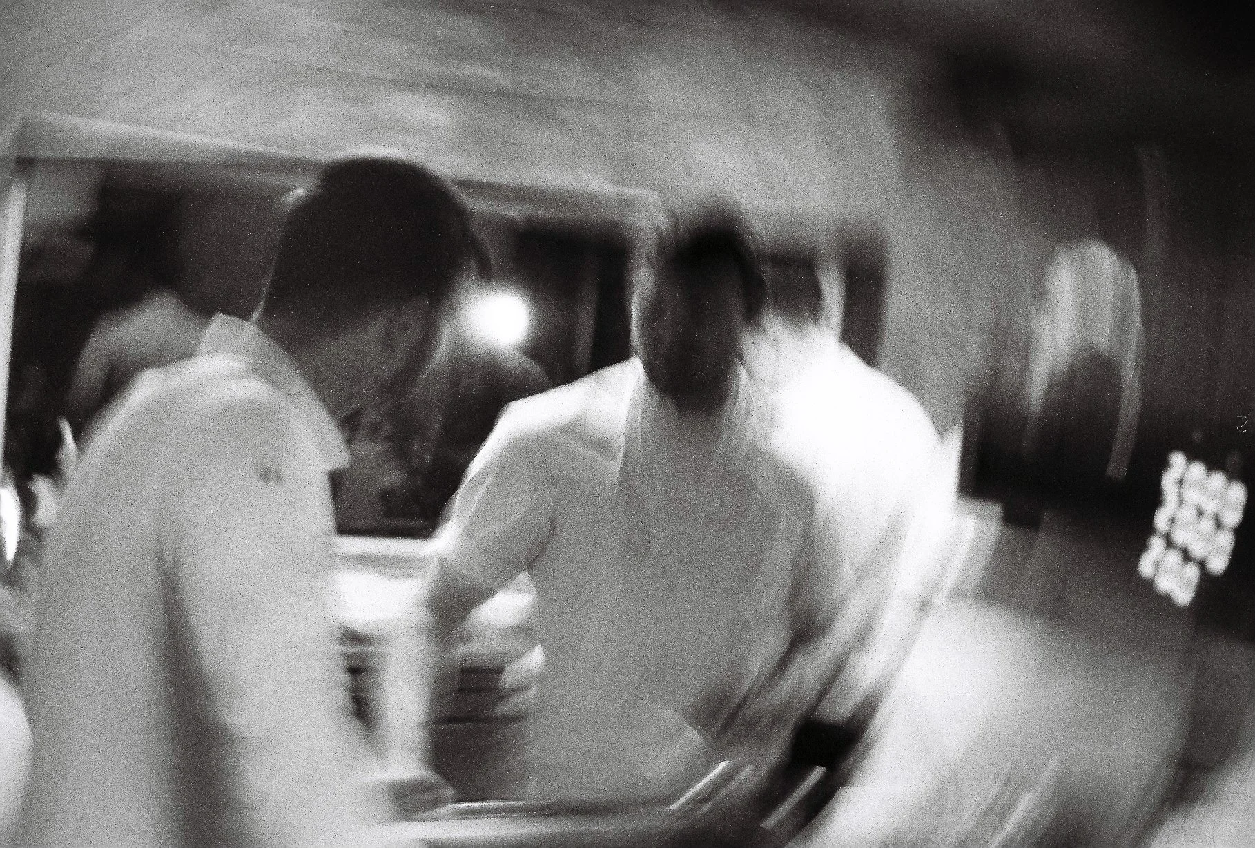 Black and white photo of three men in white shirts standing together indoors, engaged in conversation or an activity, with a mirror or reflective surface in the foreground.