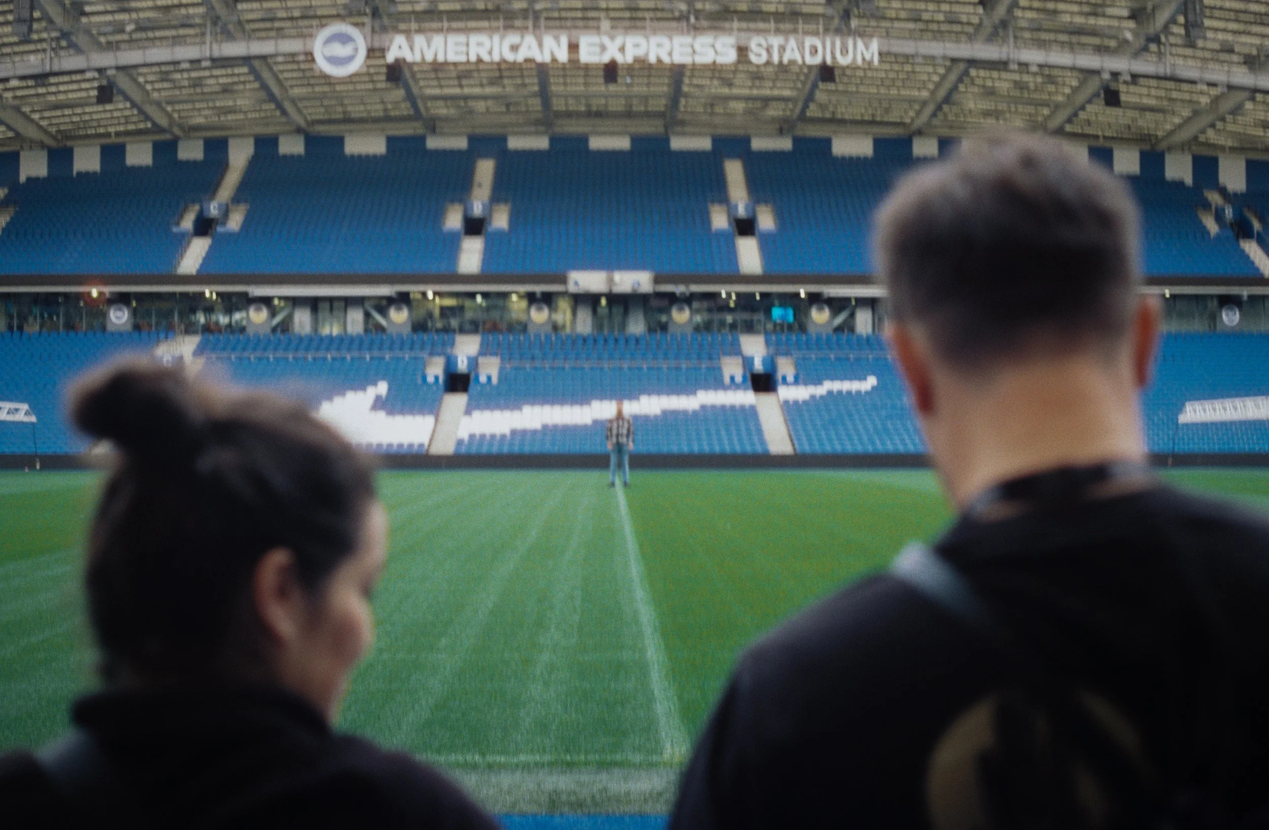 View of an empty American Express Stadium with two fans in the foreground, looking towards the field and a referee at midfield.
