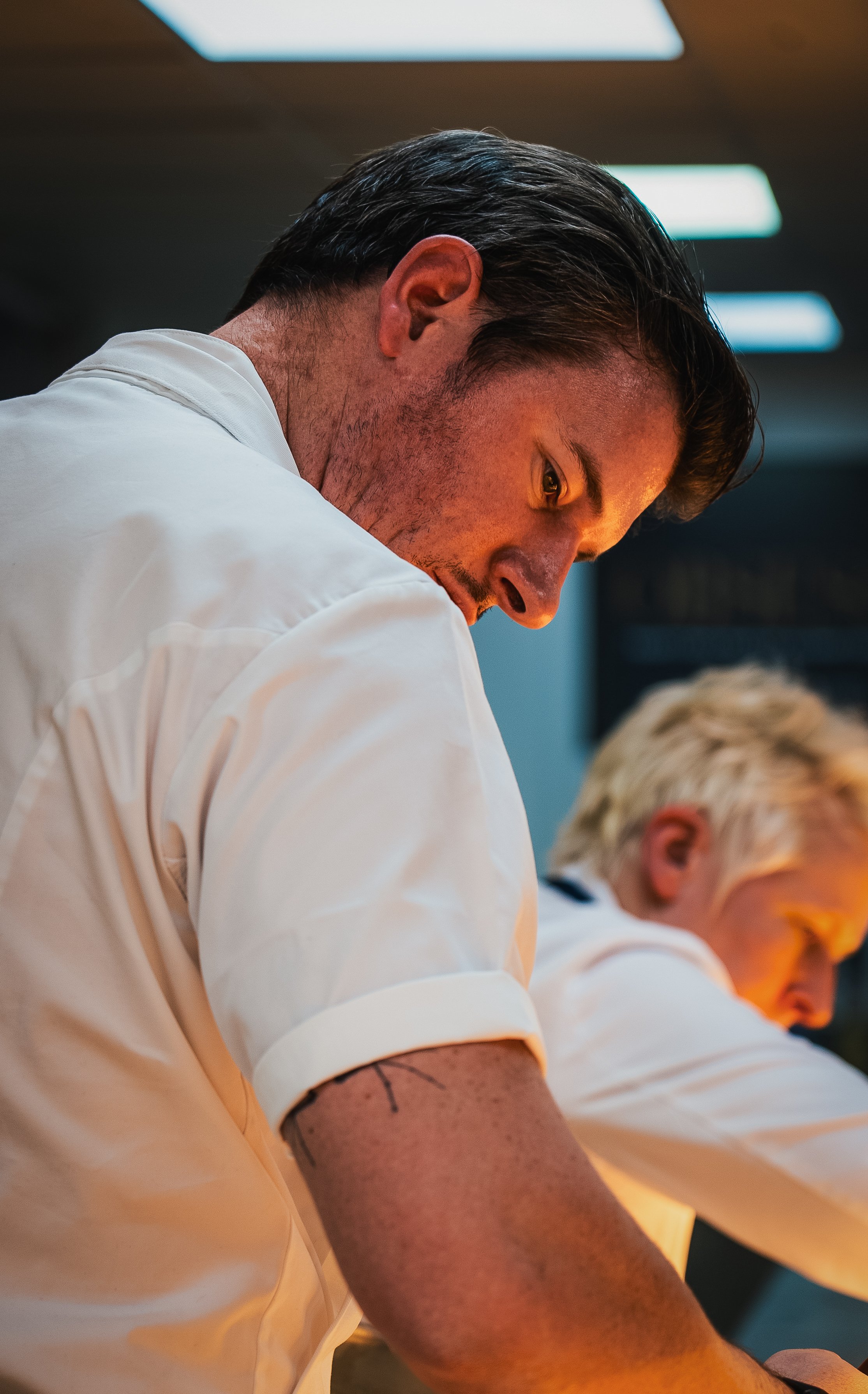 Close-up of a man with dark hair, wearing a white shirt, looking downward with a focused expression in a professional setting.