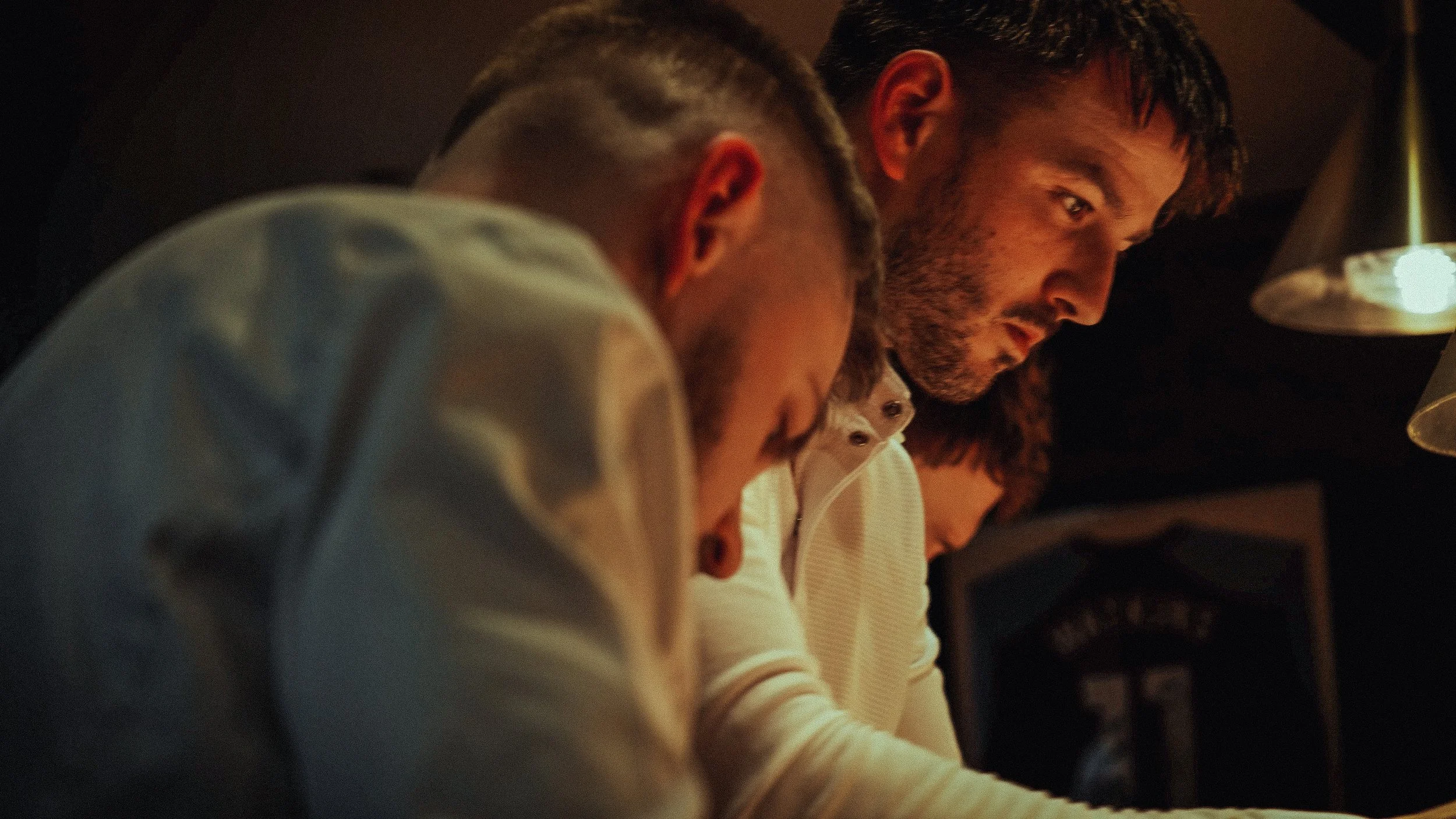 Three men sitting closely together at a dimly lit indoor setting, with their heads bowed and focused on something in front of them.