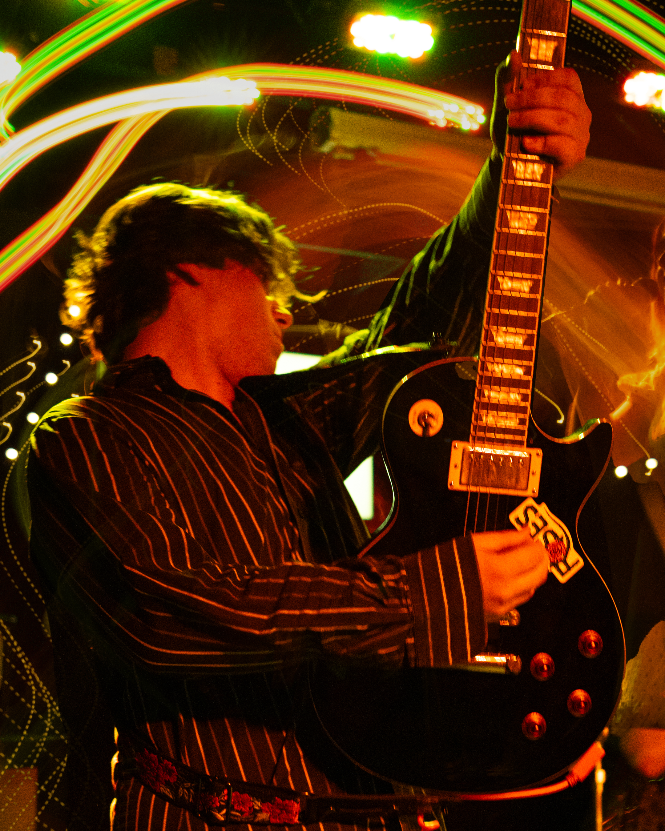 A person playing an electric guitar on stage with multicolored light trails and red lights. The musician is wearing a black and red striped shirt and has dark hair. Bright stage lighting creates a vibrant, dynamic atmosphere.