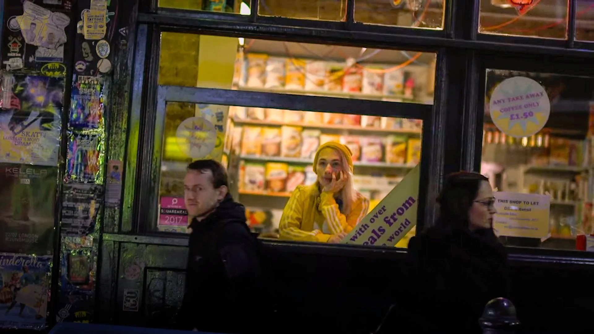 People inside and outside a small shop or kiosk, with a window displaying various items and signs. A woman in a yellow jacket and yellow headband is smiling and resting her head on her hand, while a man in a black jacket and a woman wearing glasses are sitting outside.