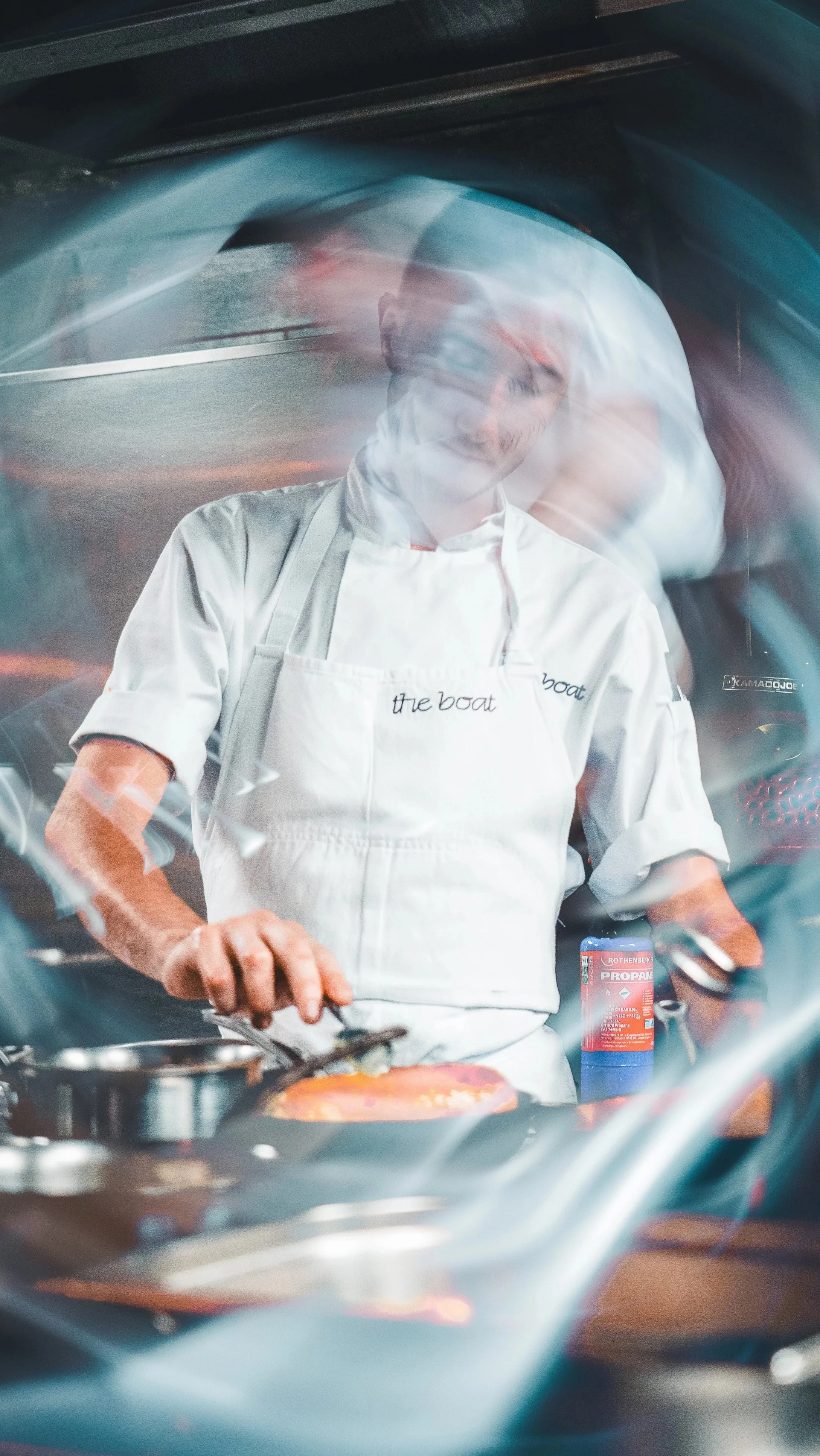 A chef in a white apron cooking on a stove with a bottle of propane nearby. The image has a motion blur effect.