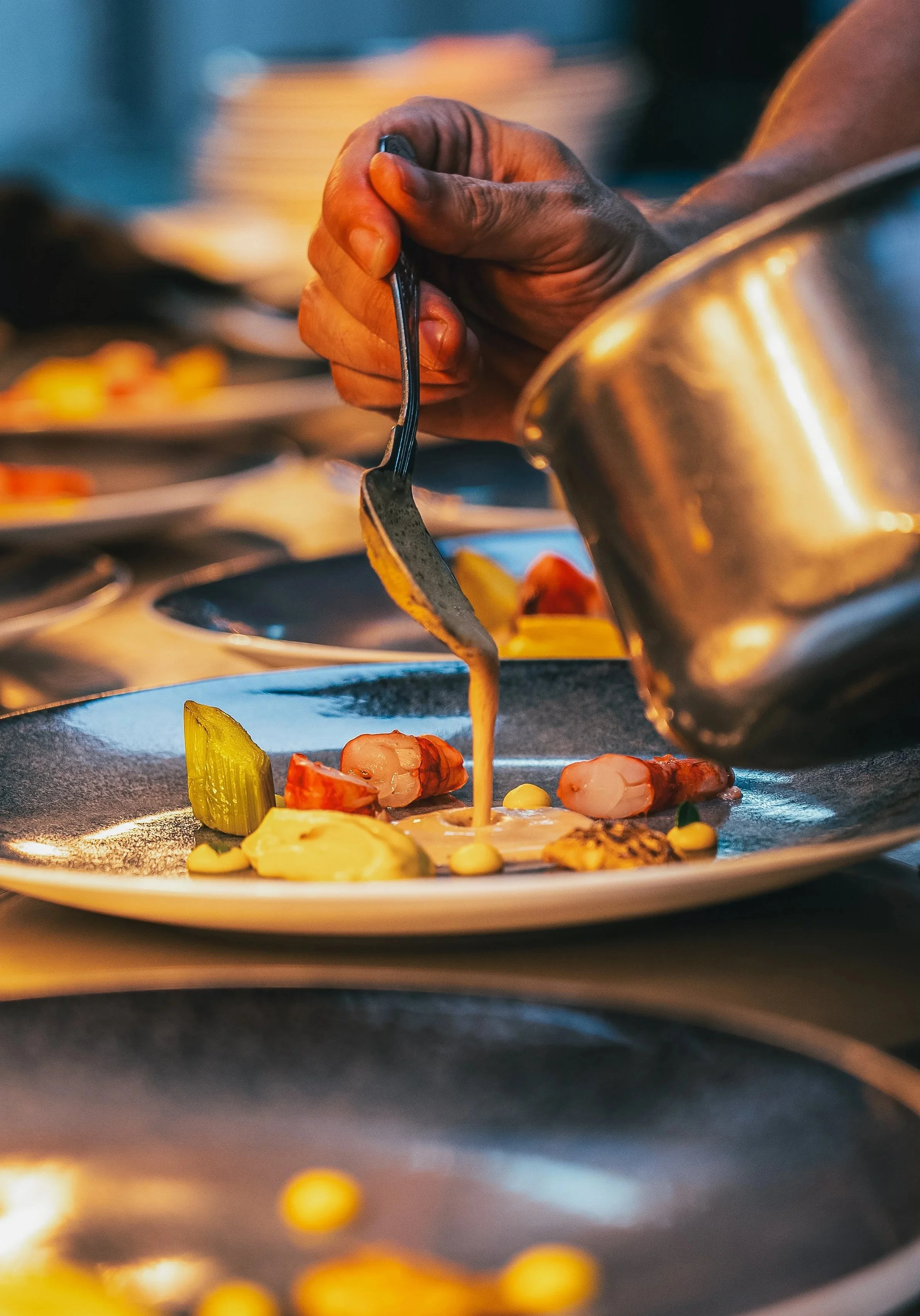 Close-up of a hand holding a fork and plating food on a dish, with various ingredients like shrimp, vegetables, and sauce visible.