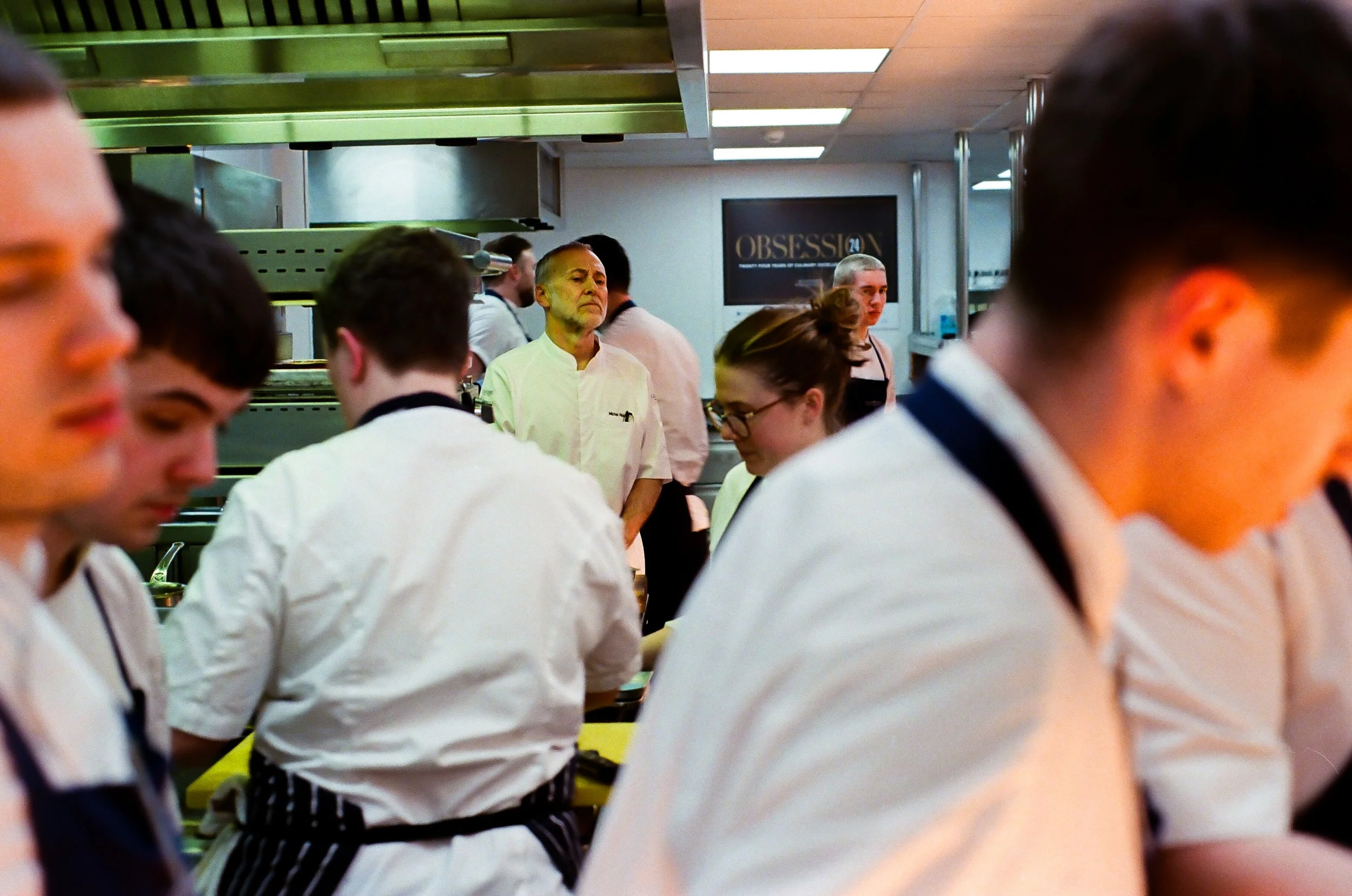 People in chef attire working in a professional kitchen with stainless steel appliances and a sign that says 'OBSESSION.'