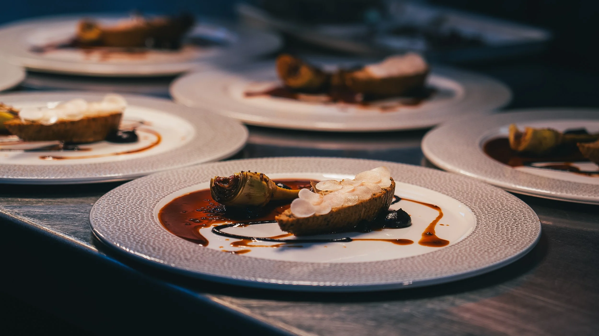 Several plates of gourmet appetizers featuring oyster shells, white sauce, and a dark sauce, arranged on a table with a blurred background.