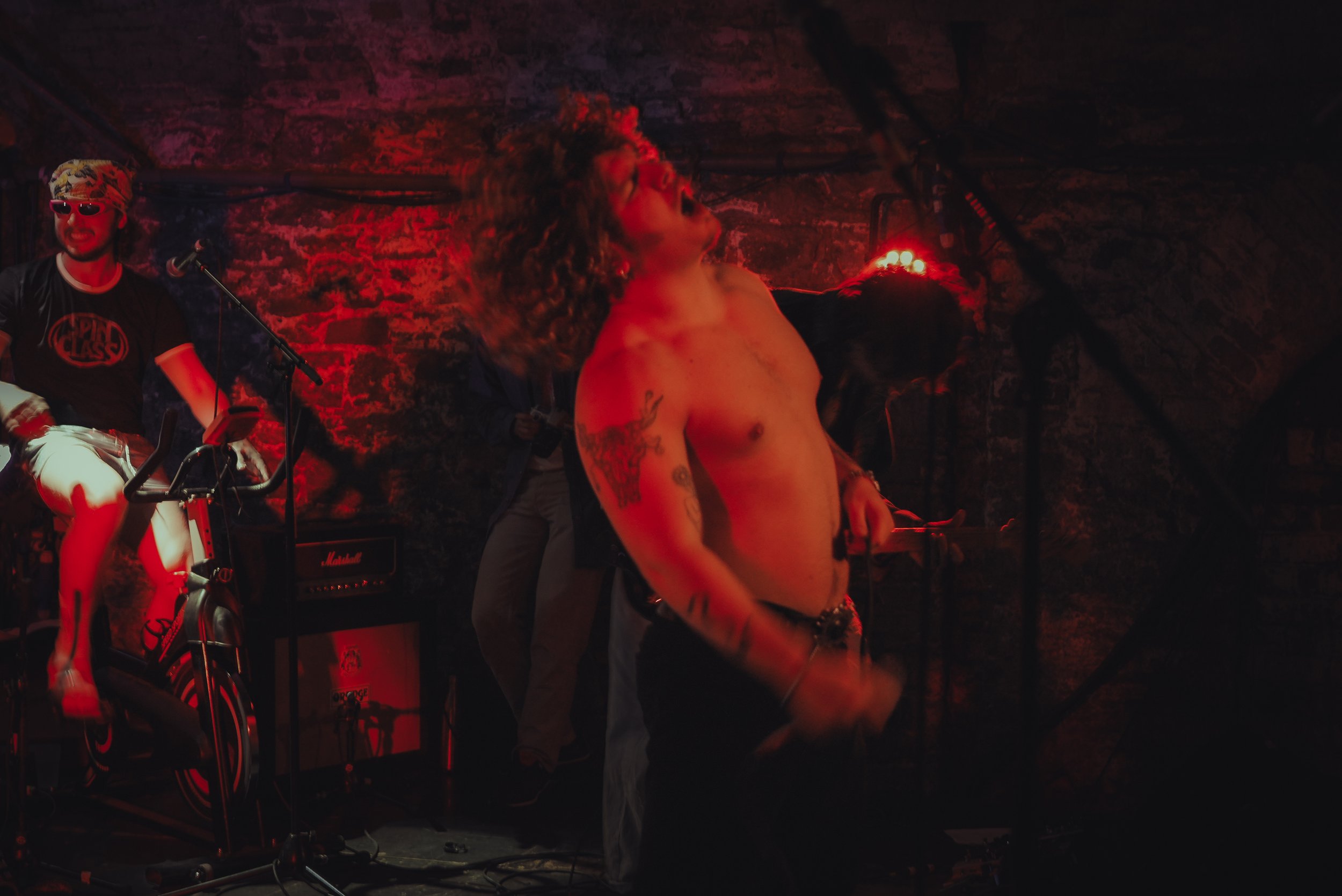 A shirtless man with tattoos on his arms and chest performing on stage, with long hair, surrounded by musicians and stage equipment, in a dimly lit environment with red lighting.