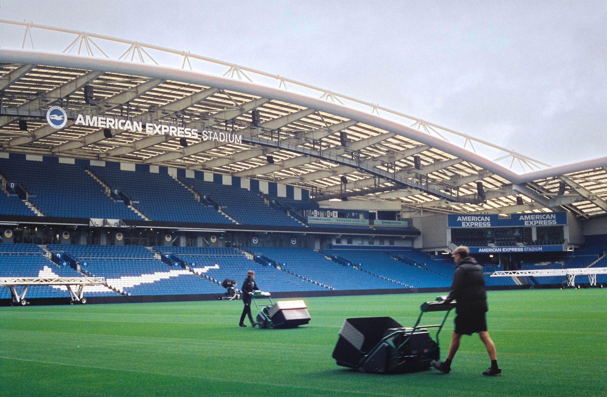 Workers preparing the field at American Express Stadium with equipment and scaffolding on the pitch for an event or maintenance, with seating and banners visible in the background.