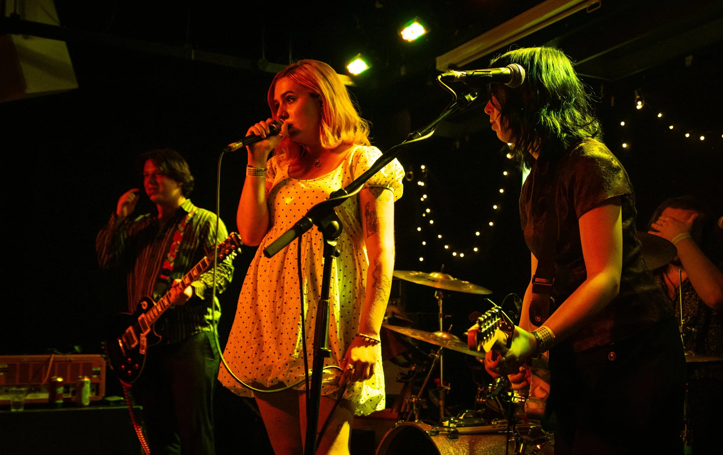 Band performing on stage with female singer in a yellow polka-dot dress holding a microphone, guitarist, and drummer in the background under dim stage lighting.