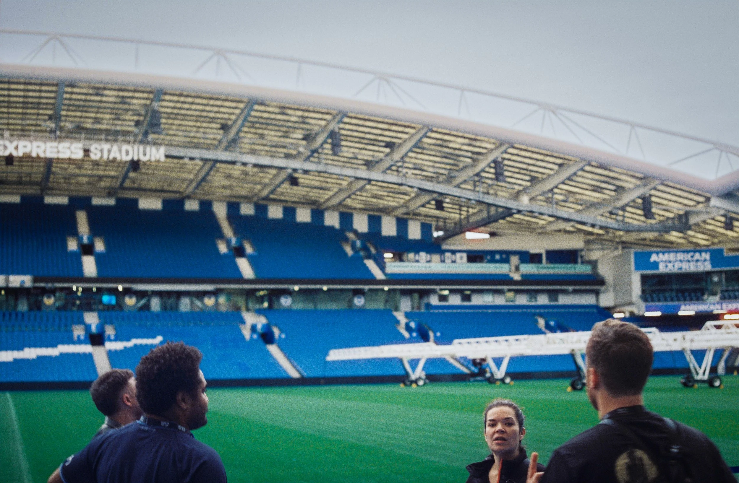 A group of people gathered on a sports field inside a large stadium with blue seating and a curved roof. The stadium is named 'EXPRESS STADIUM' with seating behind them.