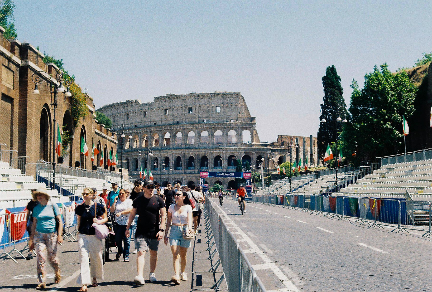 People walking and cycling in front of the Roman Colosseum in Rome, Italy, with barriers and Italian flags along the street.