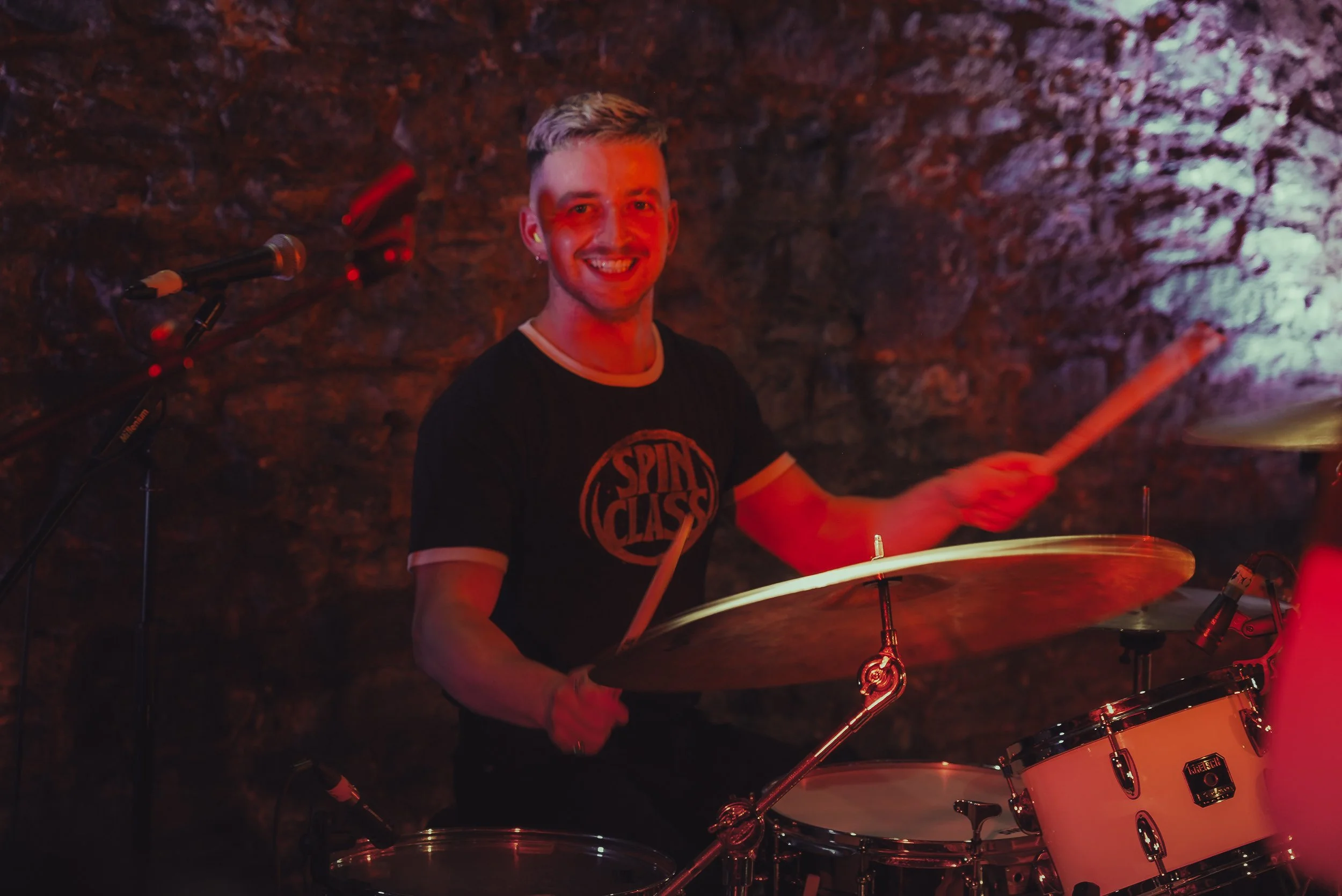 A smiling man with short blonde hair playing drums in a dimly lit setting with a rocky wall background.