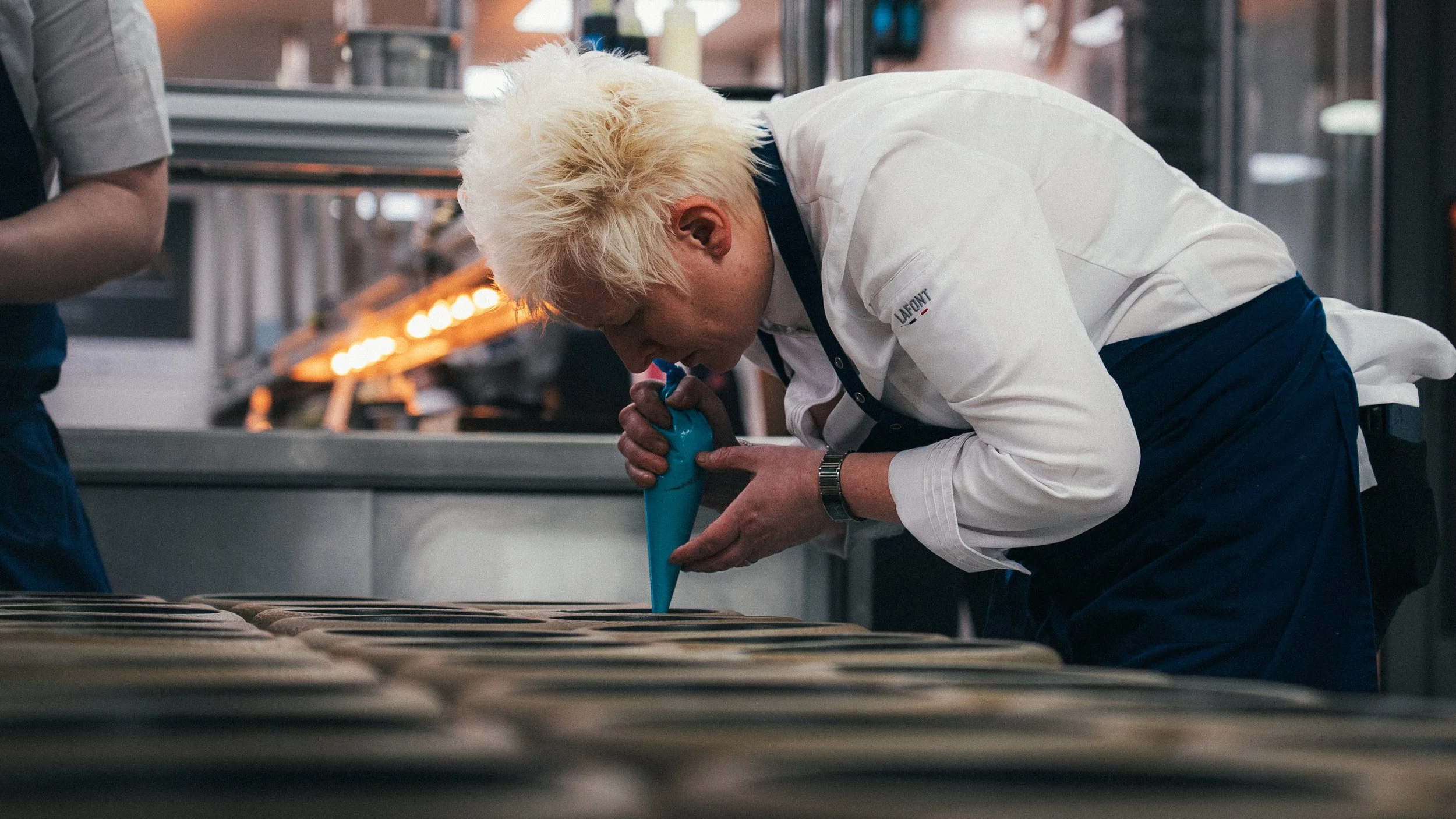 A chef with platinum blonde hair and a white chef coat is piping icing onto pastries in a professional kitchen.