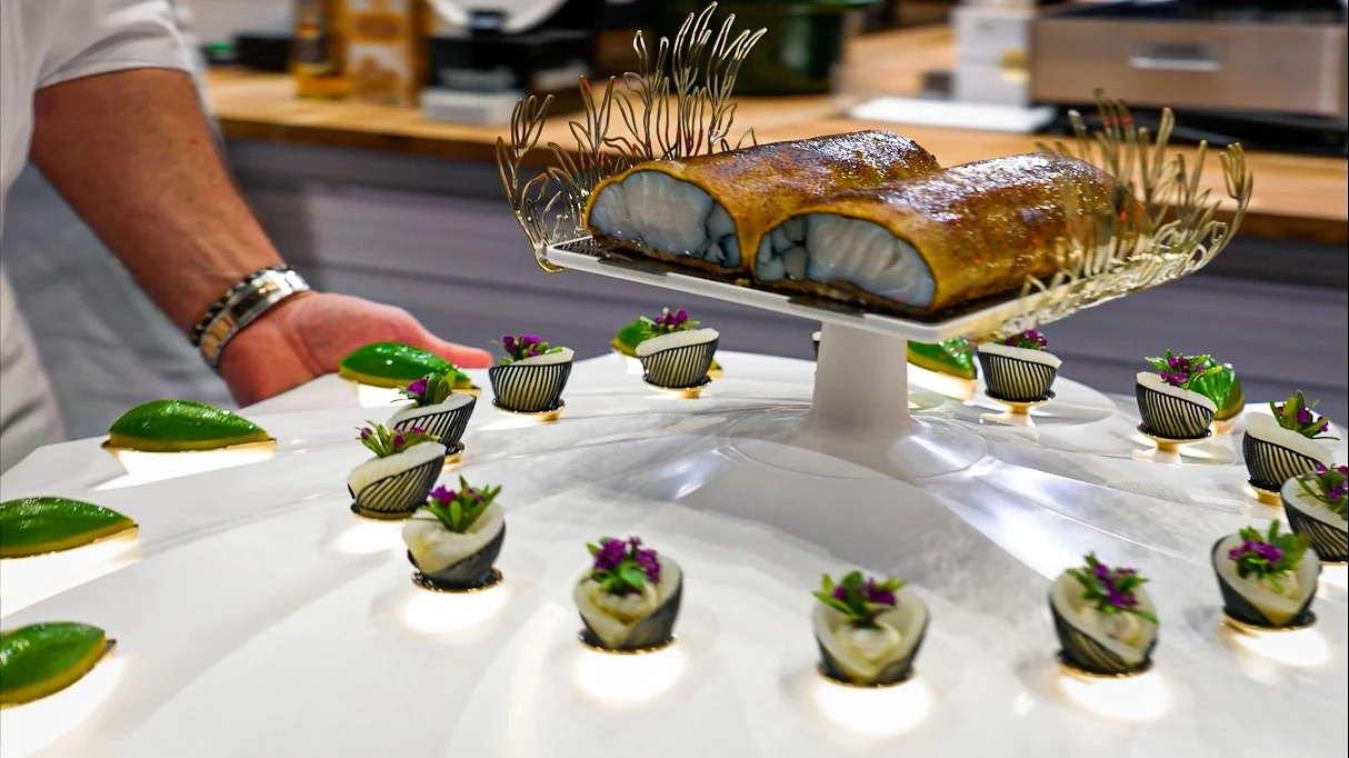 A chef presents a cooked fish fillet on a decorative serving platter surrounded by small garnished dishes and green-topped appetizers.