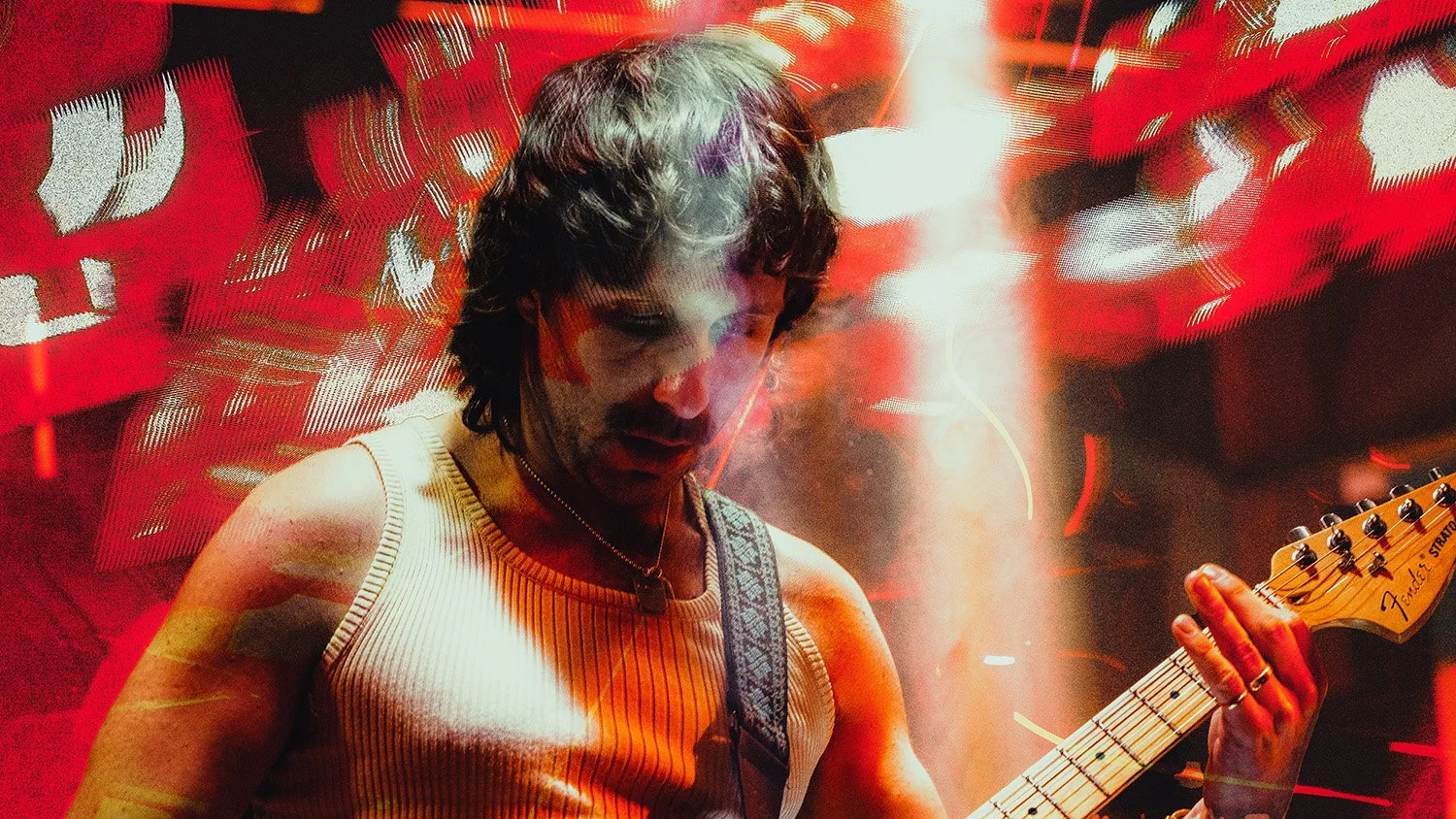 A man with dark, shoulder-length hair playing an electric guitar on stage, wearing a sleeveless, striped shirt surrounded by dynamic red and orange lighting effects.