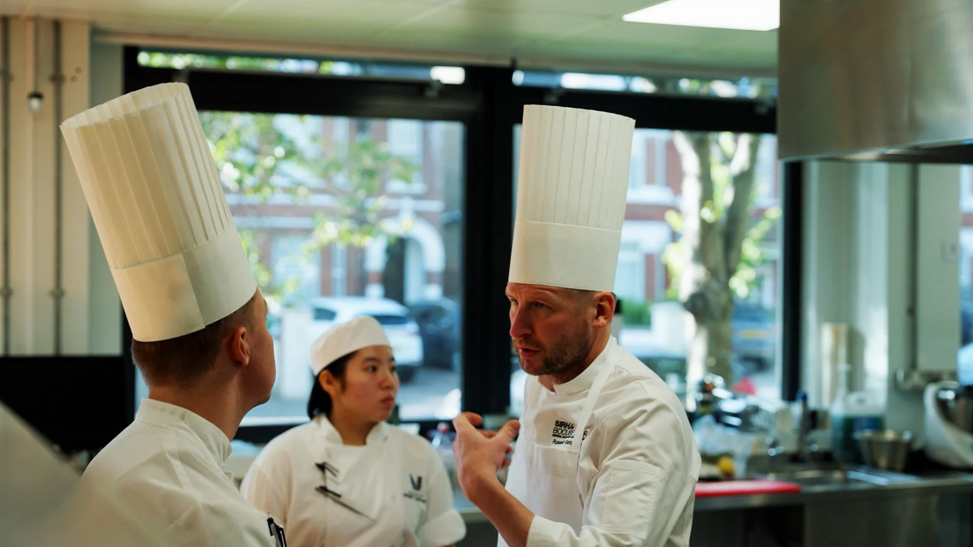 Three chefs in a kitchen wearing tall white chef hats and white uniforms, engaged in conversation.