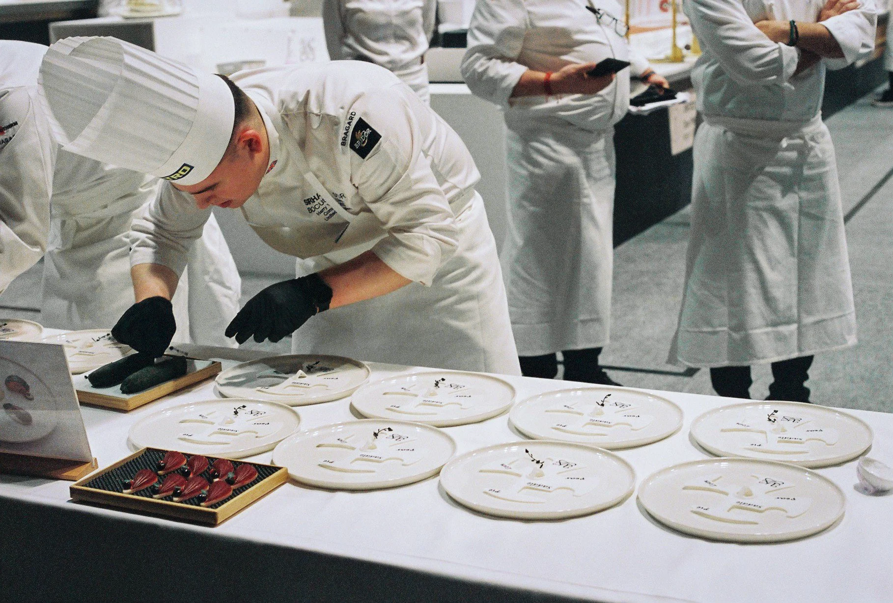 Chef in white uniform and hat working at a table with multiple plates of sliced black fish and red garnishes, while three individuals in white uniforms observe.