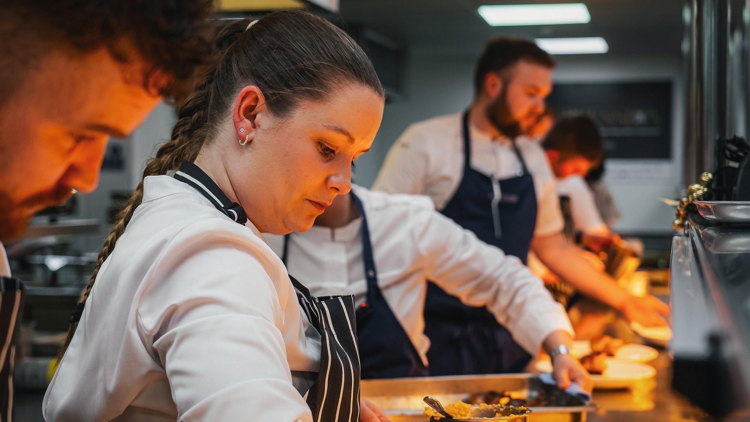 Young woman with braided hair and earrings working in a professional kitchen, preparing food alongside other chefs.