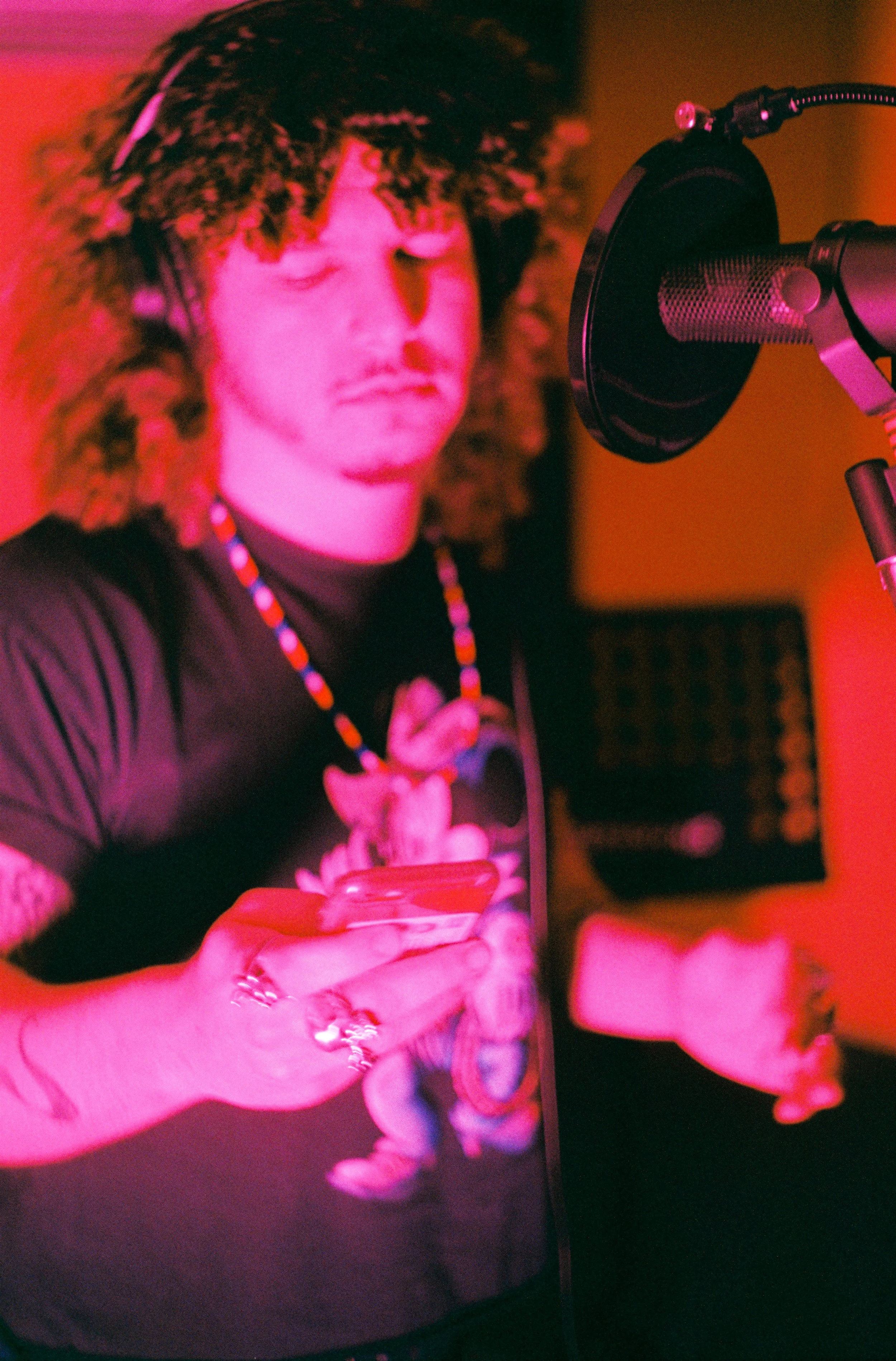 A person with curly hair wearing a beaded necklace, recording vocals in a music studio with a microphone, illuminated by pink lighting.