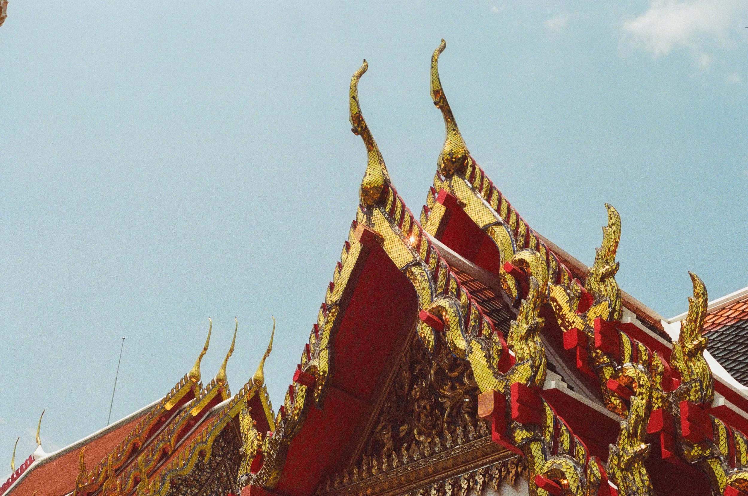 Ornate rooftop of a traditional Thai temple with golden decorative elements and a red structure, against a blue sky.