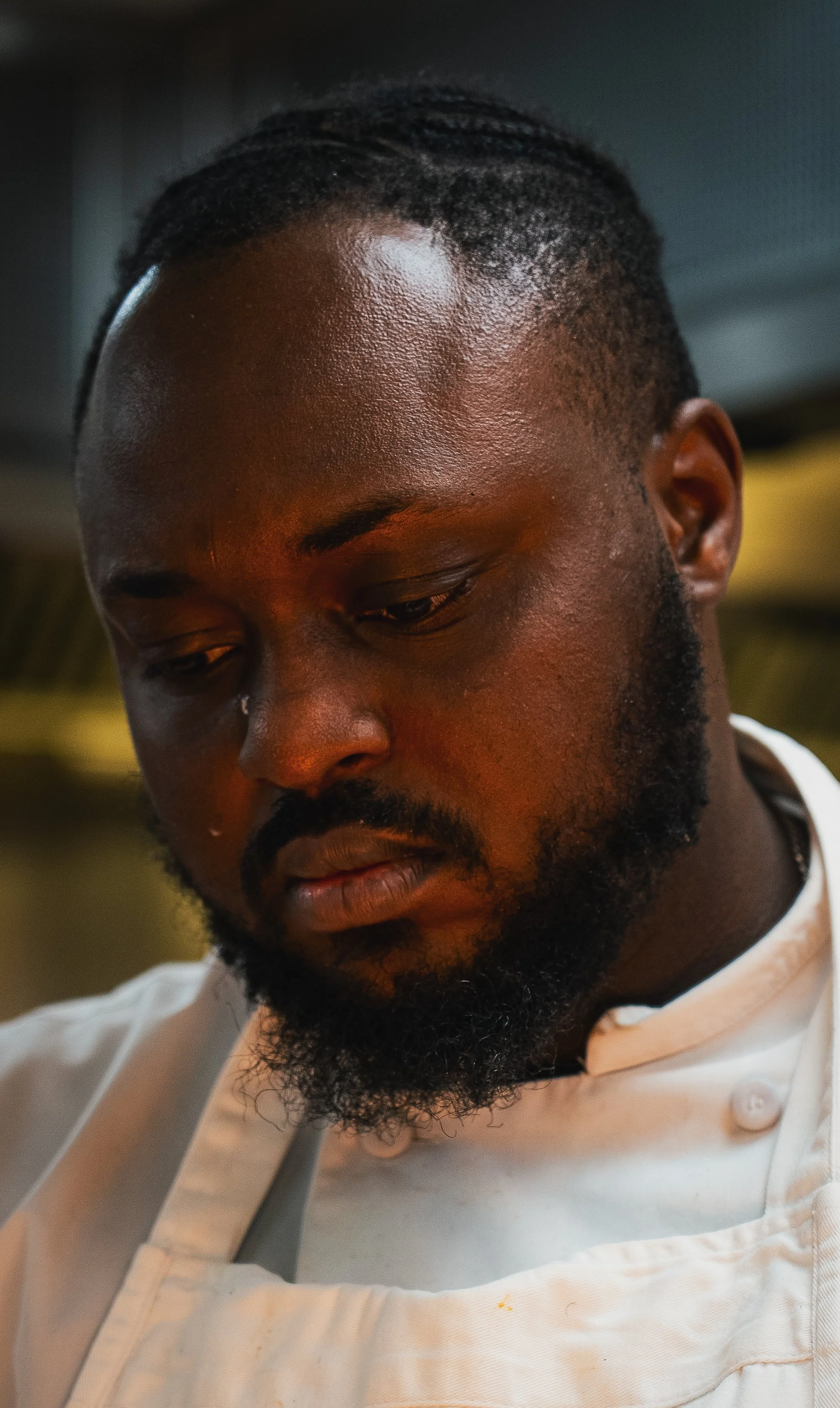 Close-up of a man with dark skin, a beard, and short curly hair, looking down, wearing a white chef's coat.