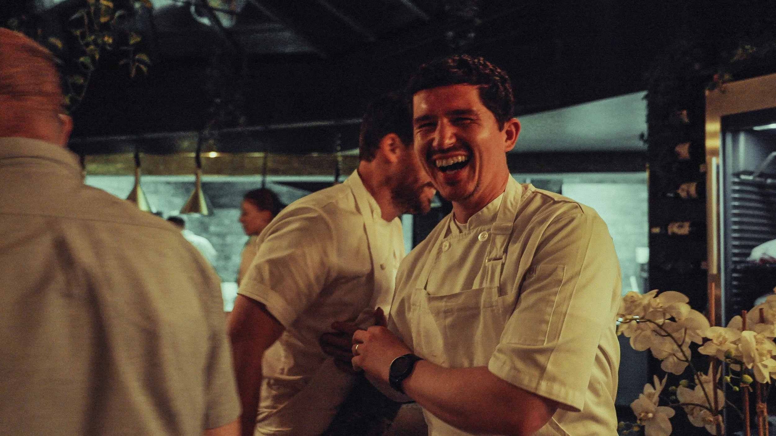 Two male chefs in white uniforms laughing and embrace in a busy kitchen with other staff in the background.