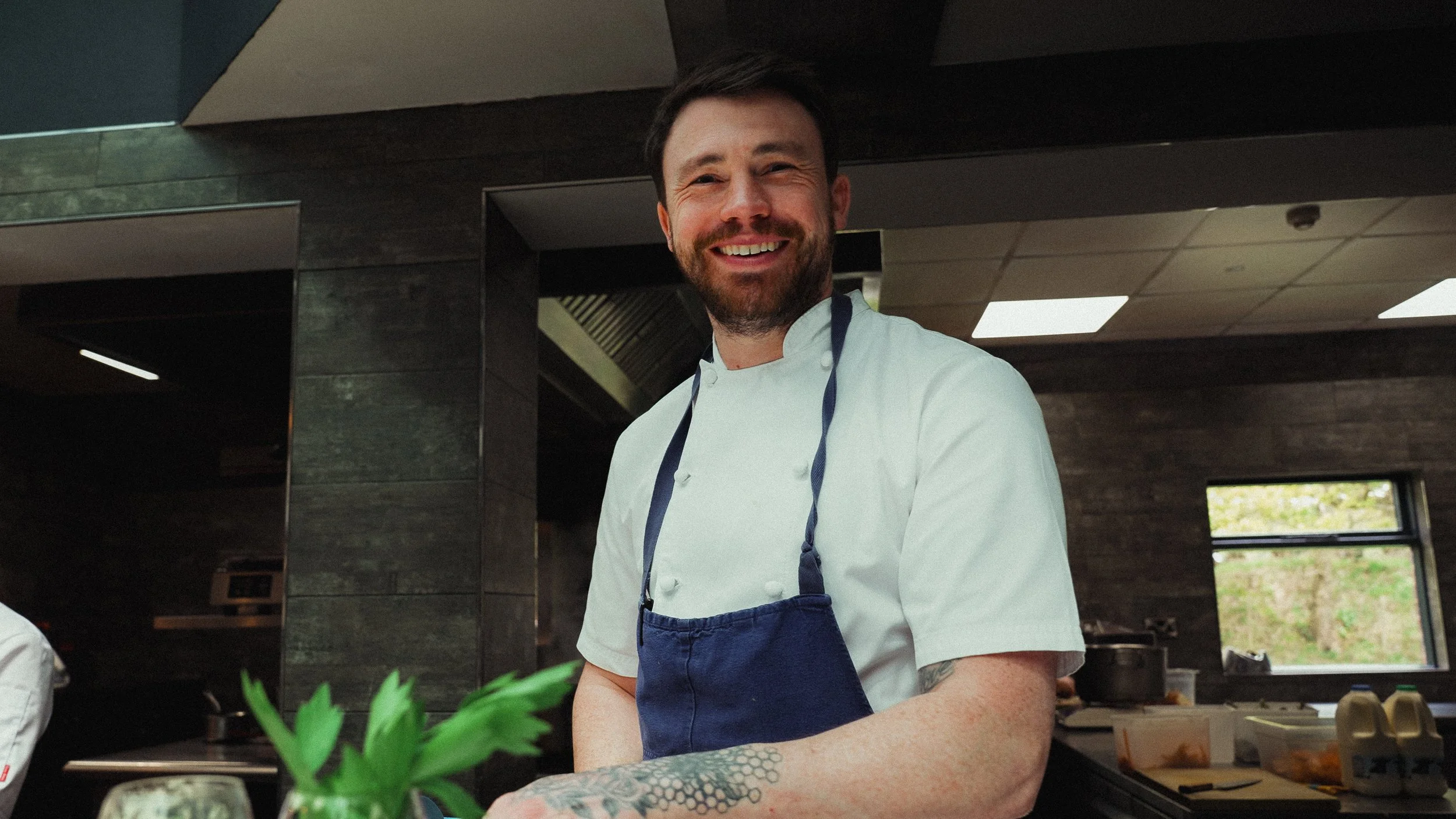 Smiling male chef wearing a white chef's coat and navy apron in a modern kitchen.