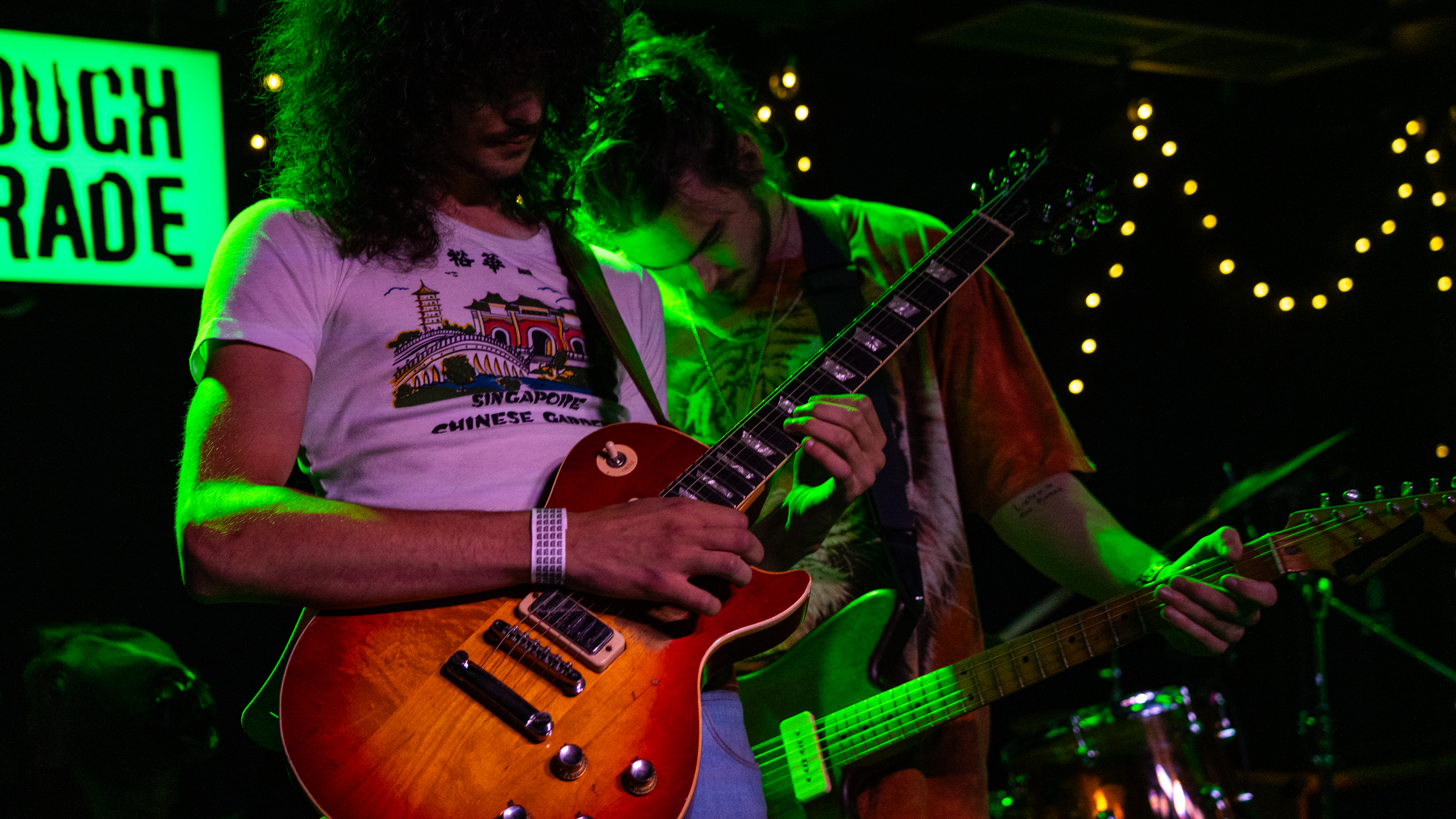 Two musicians playing electric guitars on stage under green lighting, with a sign that reads 'TOUGH TRADE' in the background.