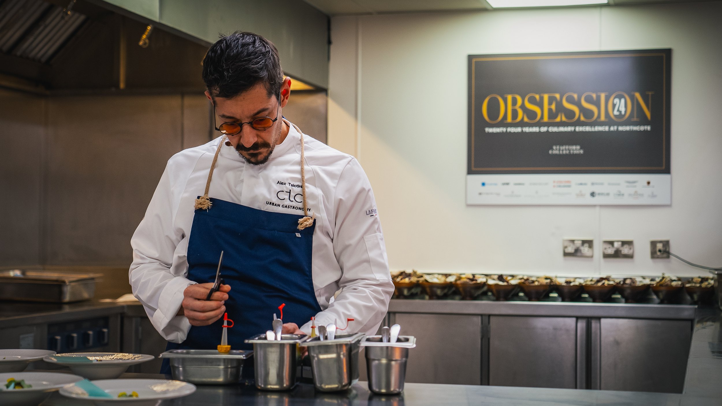 A chef wearing glasses and a white apron with blue accents is working at a kitchen counter, preparing food. Behind him on the wall, there is a large poster that reads 'OBSESSION 24' and mentions twenty-four years of culinary excellence at Northcote.