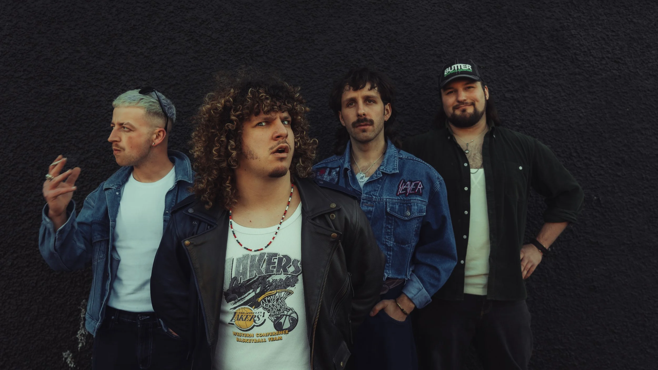 Four young men standing against a black textured wall. They are dressed in casual clothing, with two wearing denim jackets, one in a leather jacket, and one in a black shirt. One man has light hair dyed blue, and another is wearing a cap. They appear