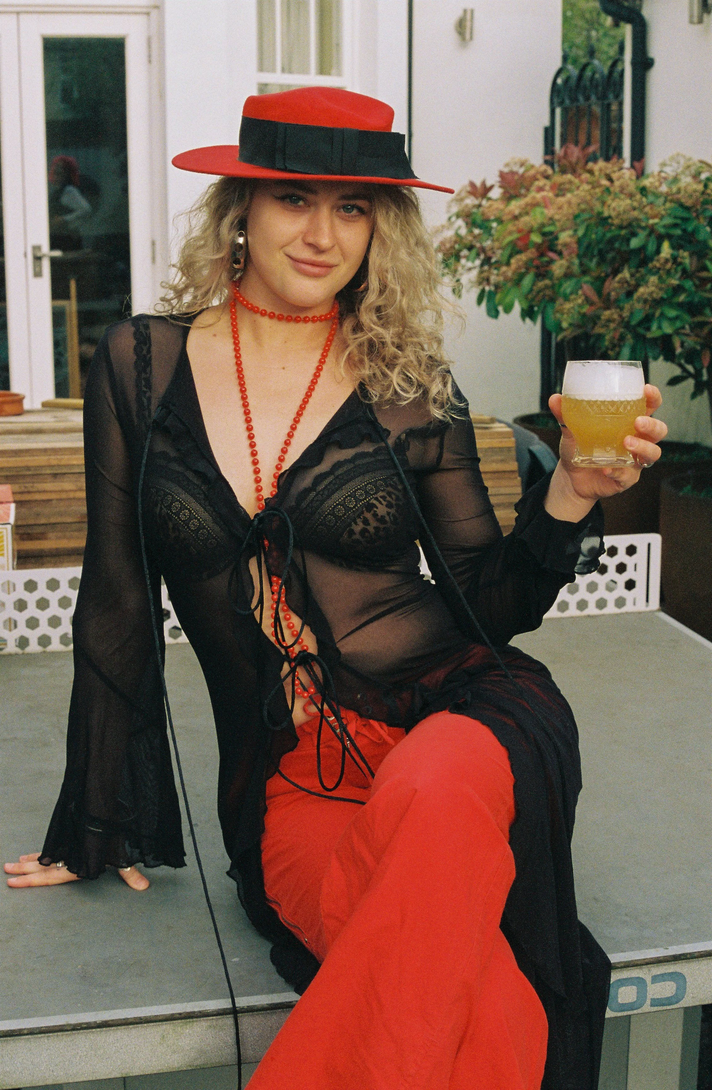 A woman with curly blonde hair wearing a red hat with a black band, a black sheer lace top, red beaded jewelry, and red pants, sitting by a mirror. She is holding a glass of beer and smiling.