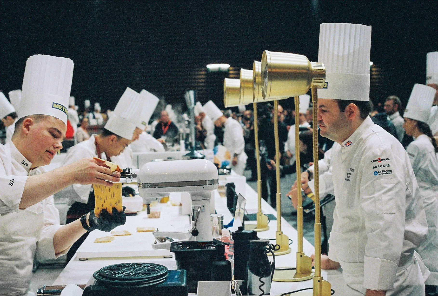Chefs working at a culinary competition station with various equipment and ingredients, with a crowd of spectators in the background.
