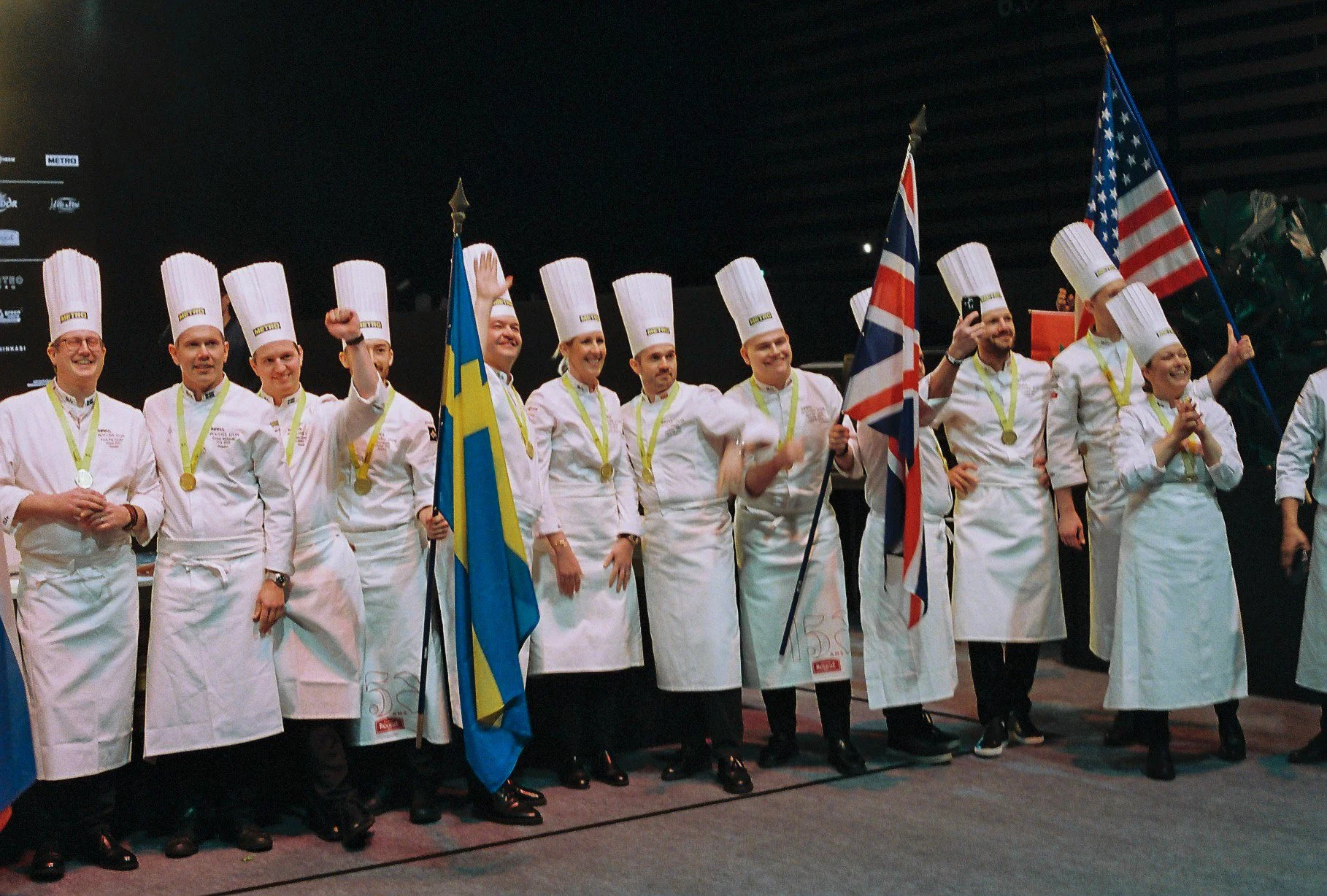 Group of chefs in white uniforms and tall chef hats, some holding flags, standing on stage with medals around their necks, celebrating at an event.