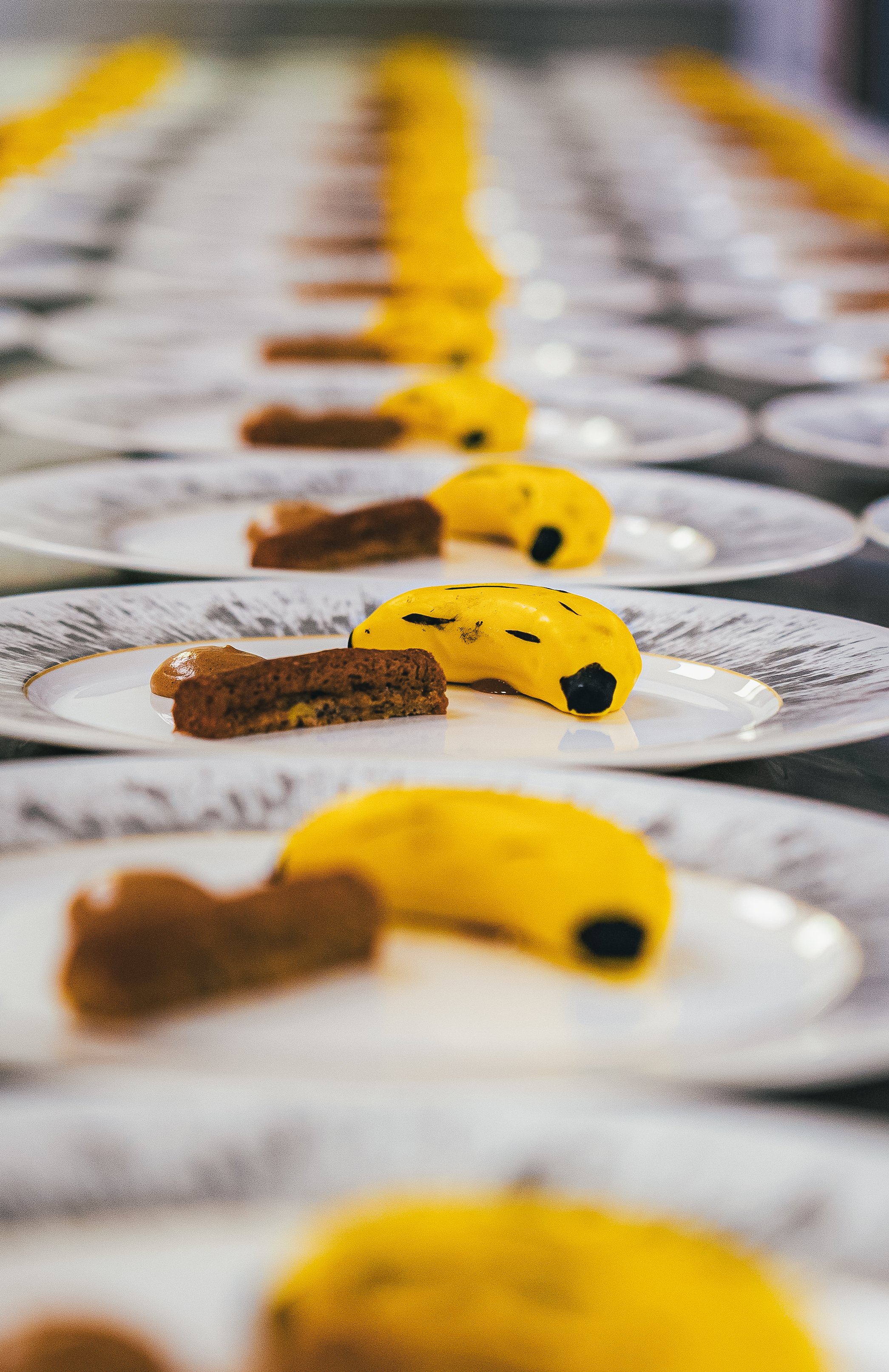 Rows of plates with various desserts, including a banana-shaped yellow treat with black spots and a chocolate-colored dessert, on a catering table.