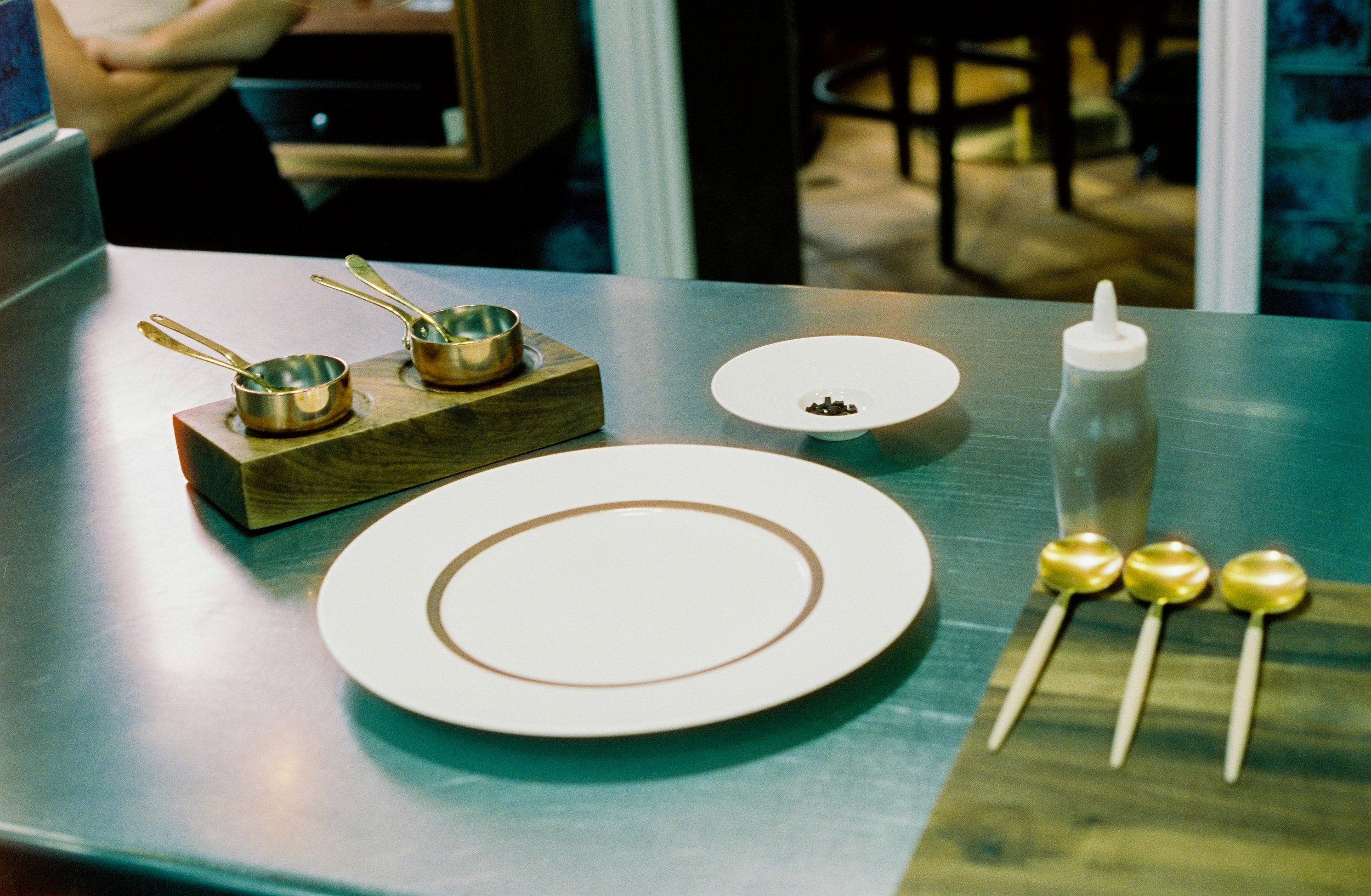 A tabletop scene with two small brass measuring cups on a wooden holder, a white plate with a circular design, small black objects in a white bowl, a white squeeze bottle, and skewers with brass-colored balls, set on a dark table in a restaurant or kitchen environment.