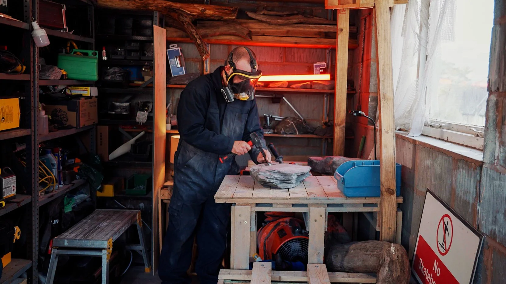 Man working with a rock on a workbench in a garage or workshop, wearing safety mask and goggles, surrounded by shelves with tools and supplies, near a window with curtains.