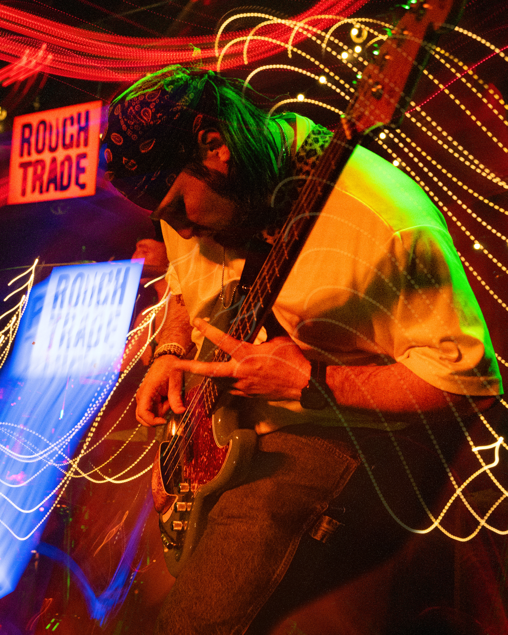 Blurred photo of two women with long hair, one with glasses, playing guitars in a dimly lit environment with colorful light trails, neon signs reading 'ROUGH TRADE' visible in the background.