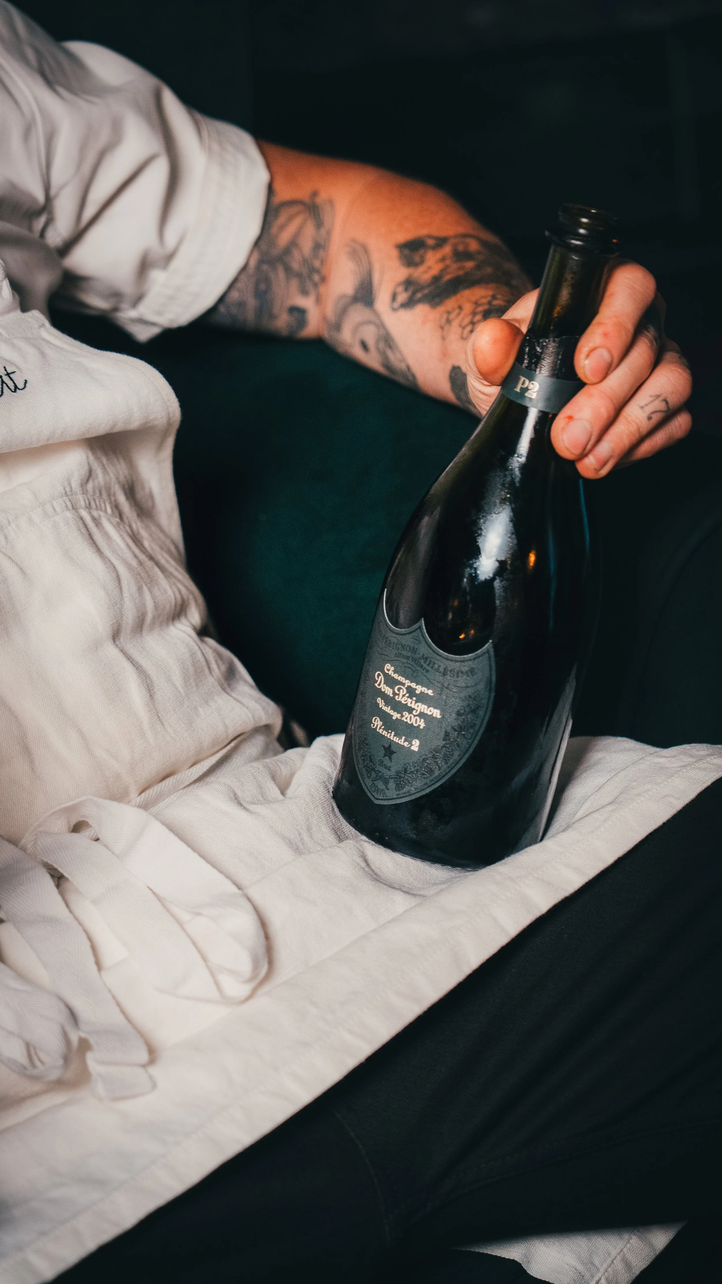 Person holding a dark bottle of Dom Pérignon champagne with tattoos on their arm, sitting on a black surface, possibly a couch. The bottle's label indicates it is vintage 2004.