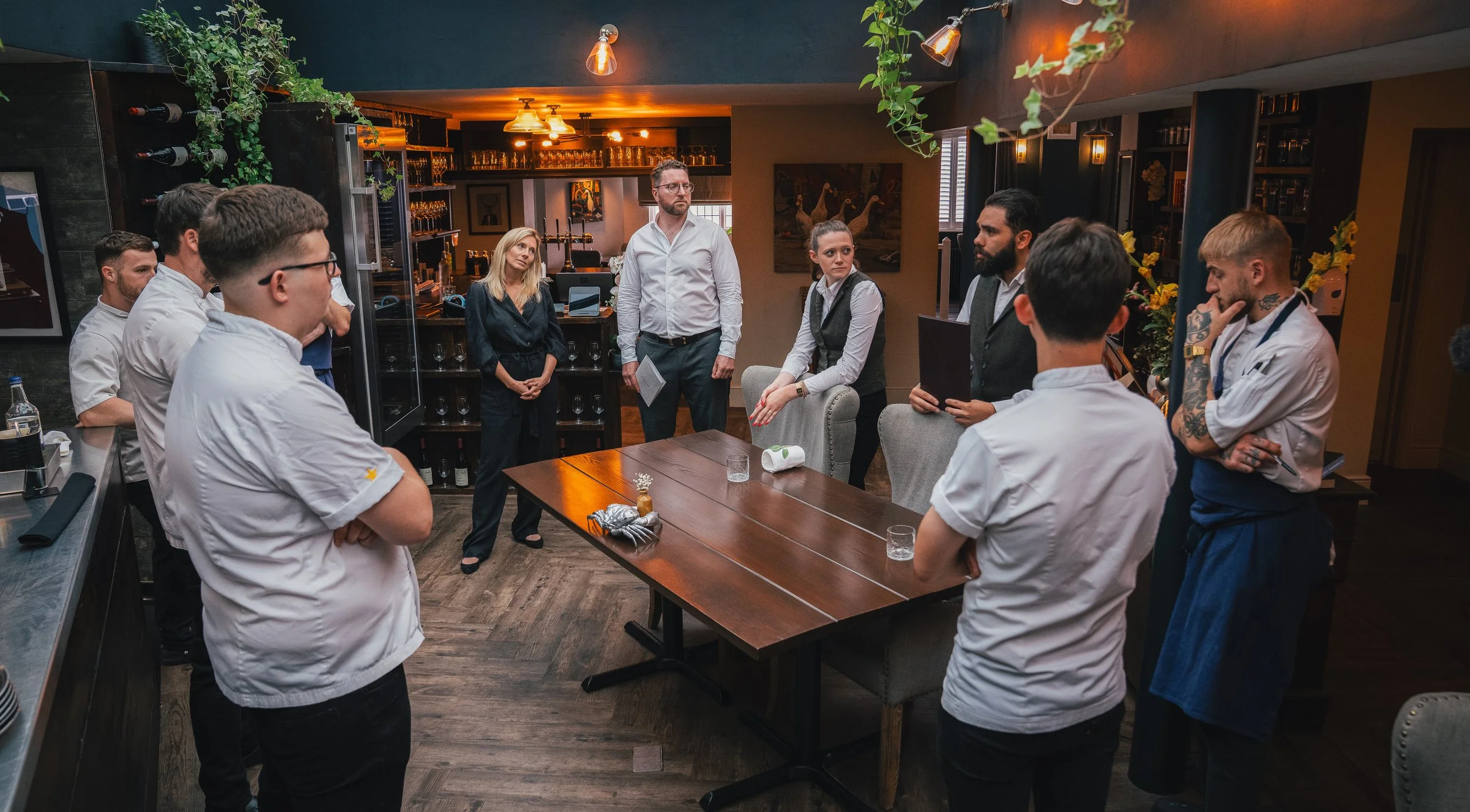 Group of chefs and staff in a restaurant kitchen/meeting area, standing around a wooden table, listening to a man speaking, with wine bottles and glassware in the background.