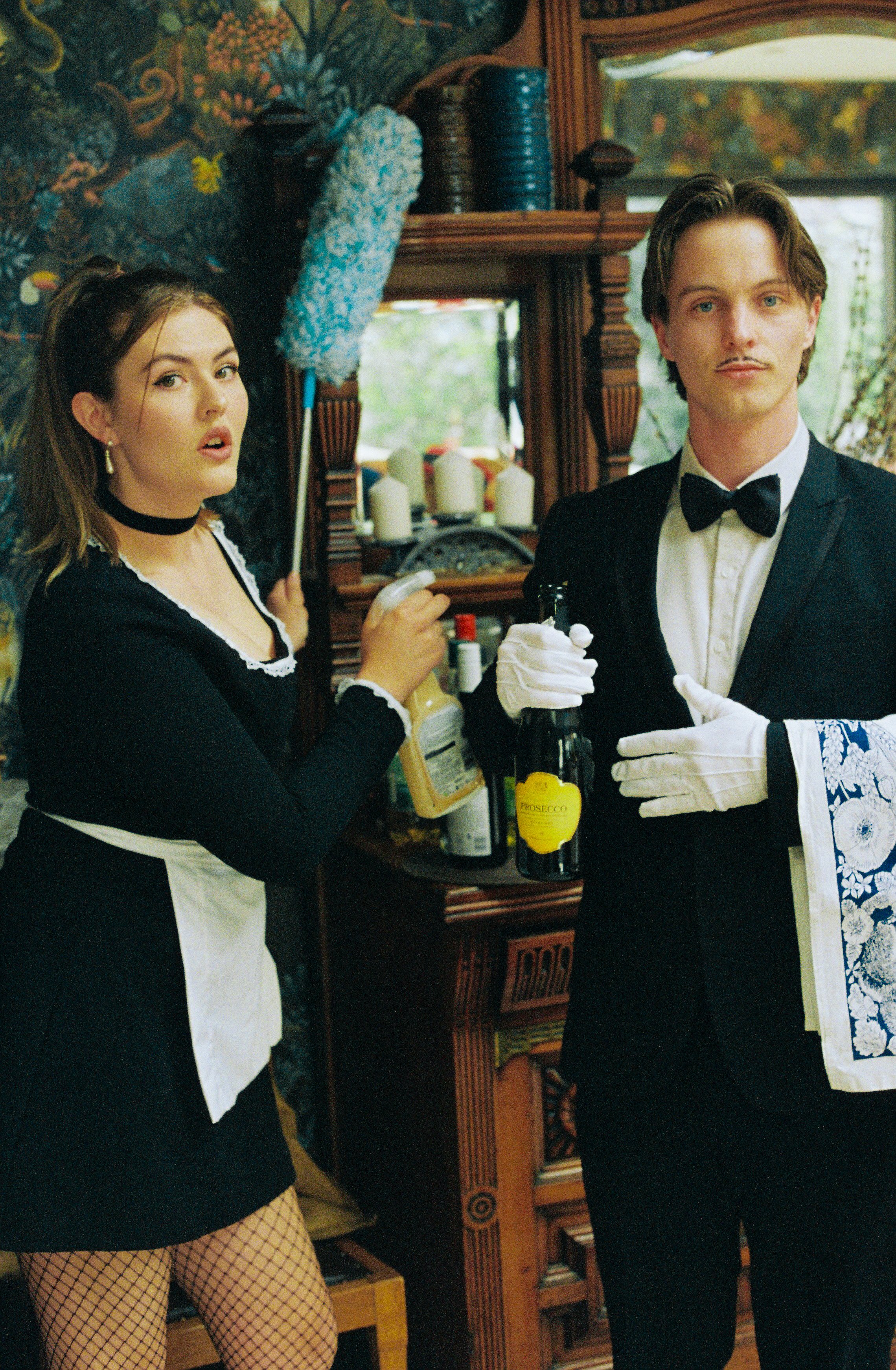 Two people dressed in costume, a woman dressed as a maid with fishnet stockings and a man in a tuxedo with fake mustache, standing in a room with a wooden corner cabinet and decorated with candles and a mural of a forest scene.