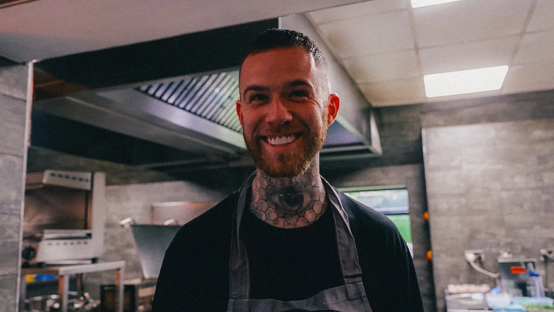 A smiling man with tattoos and short dark hair, wearing a black shirt and apron, stands in a commercial kitchen.