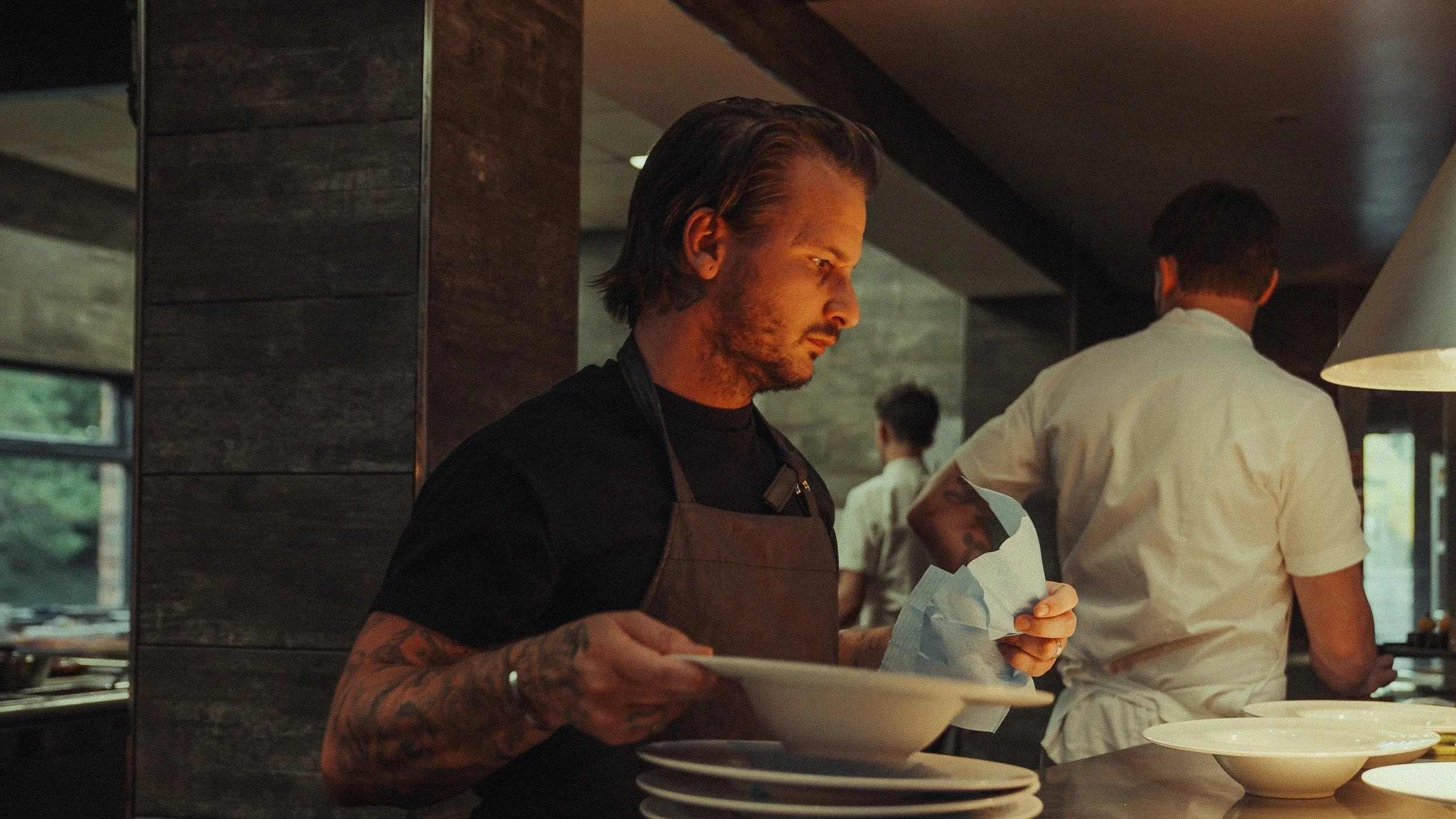 A man with long hair and tattoos wearing a black shirt and apron, standing in a restaurant kitchen, holding plates, with a chef and other staff working in the background.