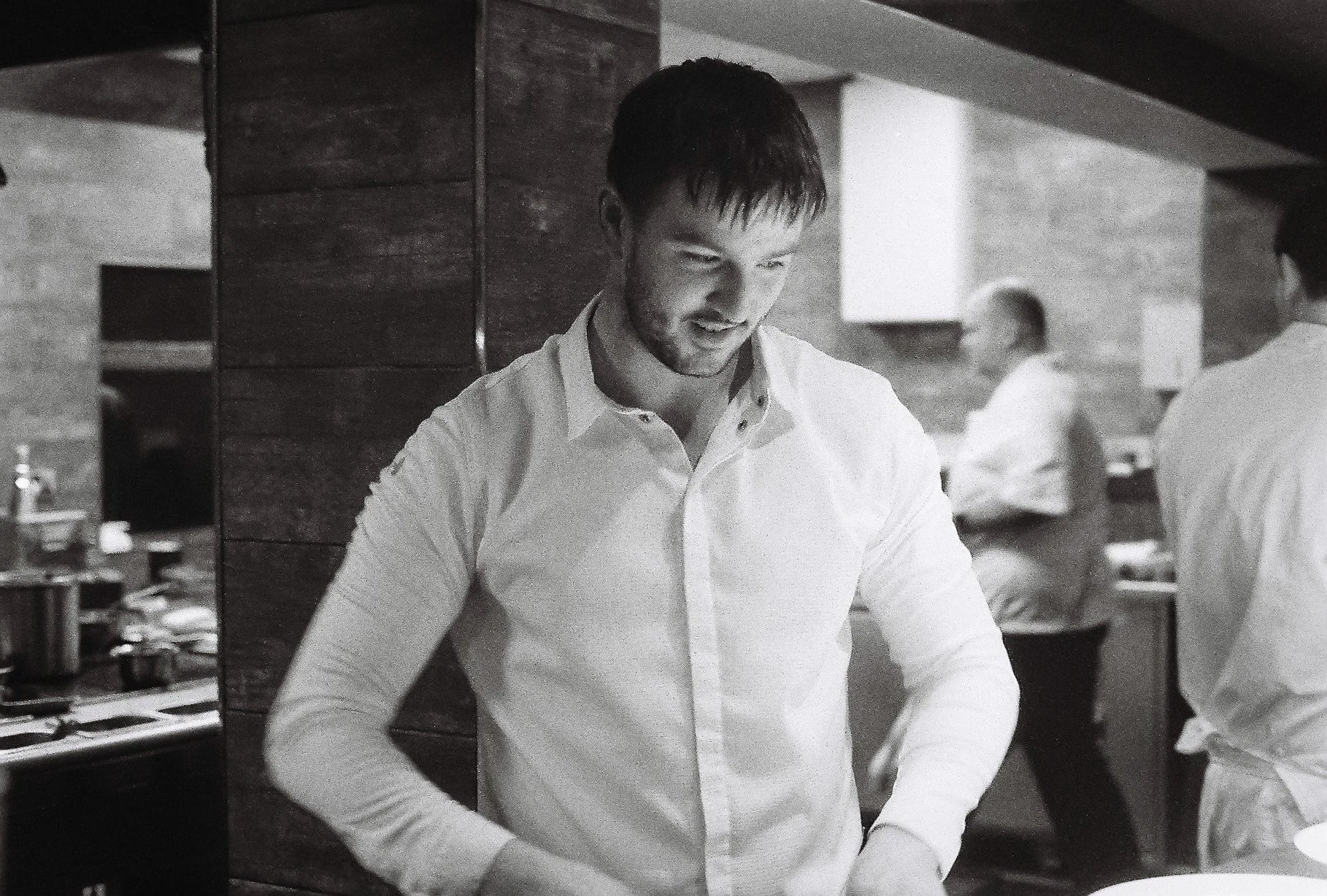 A young man with dark hair and a beard, wearing a white button-down shirt, is standing in a restaurant or cafe. He appears to be preparing or serving food, with a focused expression on his face. In the background, there are other people, including a woman in a sleeveless top, and items like plates and kitchen equipment are visible.