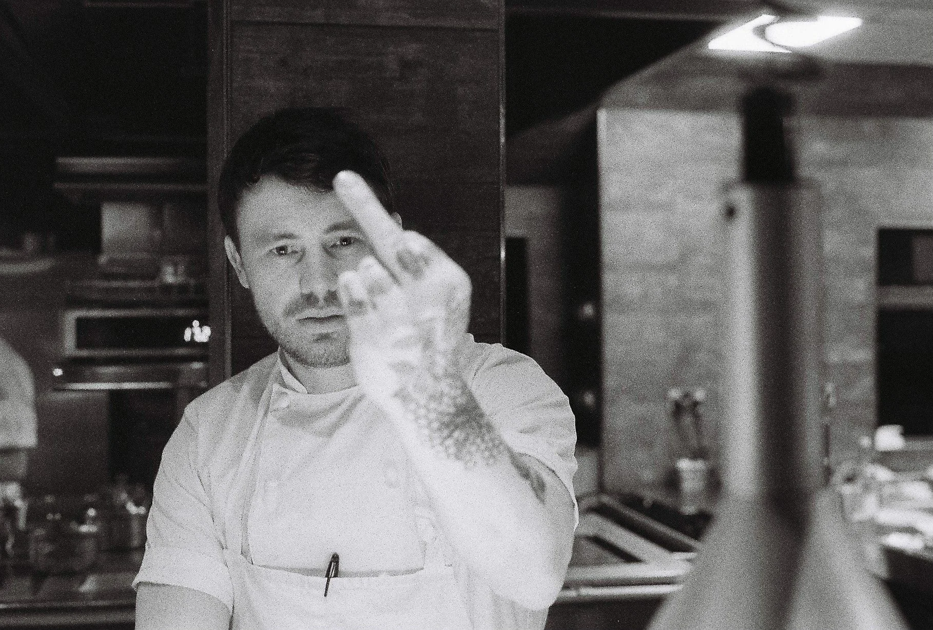A man in a kitchen wearing a white chef's coat, making an obscene gesture with his middle finger raised.